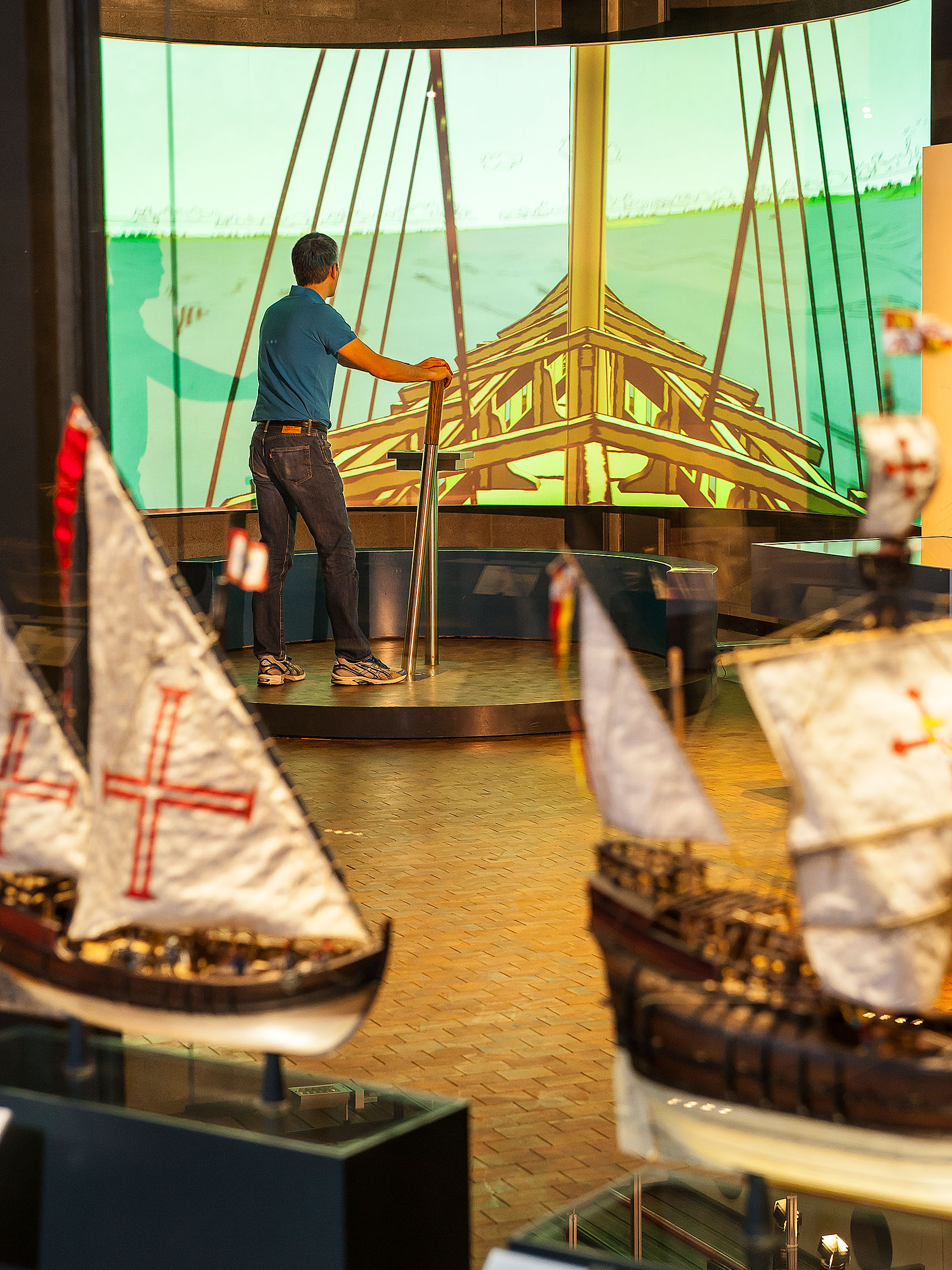 Ein Mann steht auf einem Podest und steuert ein Segelschiff, das vor ihm auf die Wand projiziert wird.