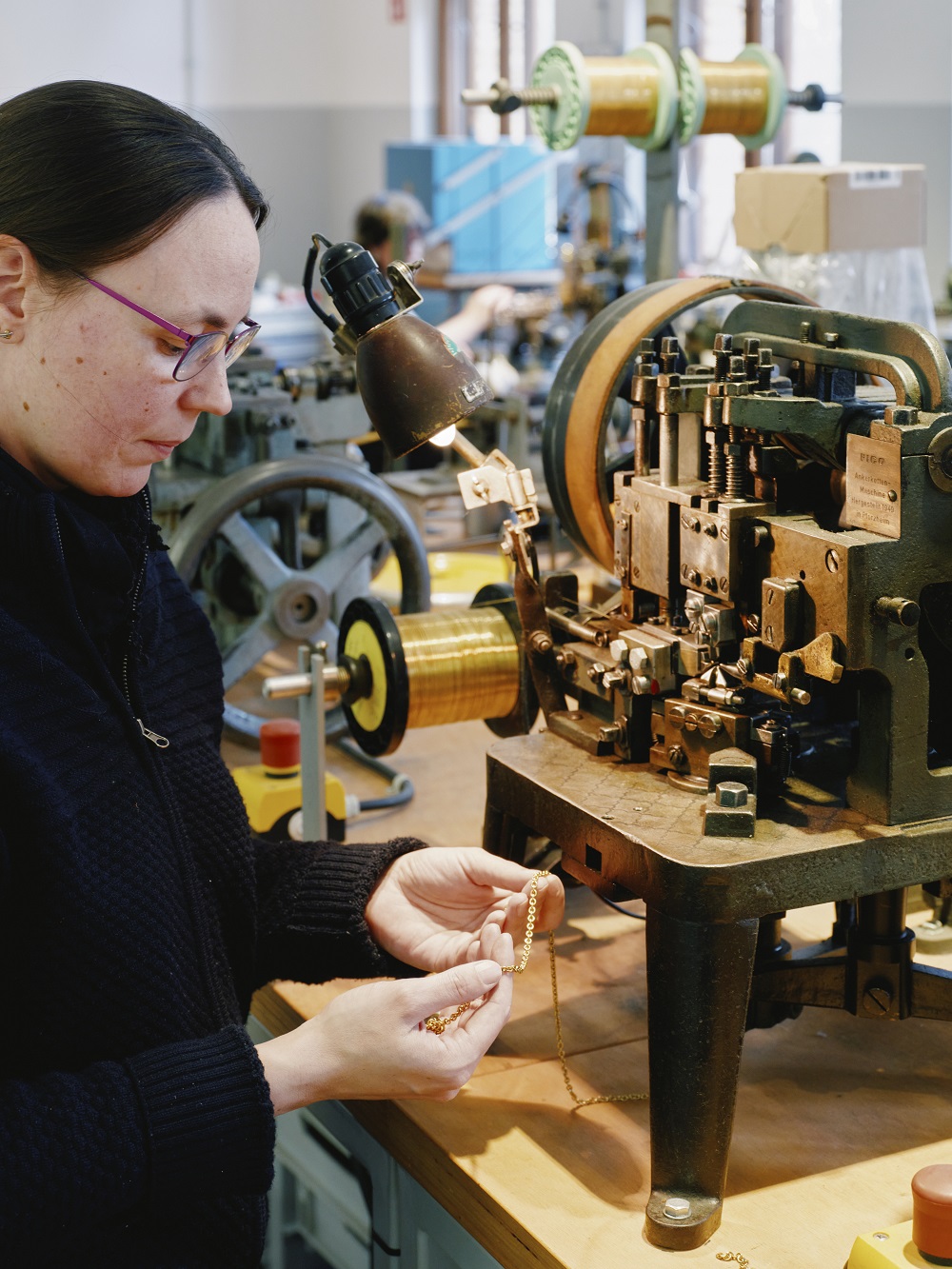Eine Frau hält eine Goldkette in der Hand. Vor ihr eine Maschine, die zur Produktion dieser Ketten verwendet wird.