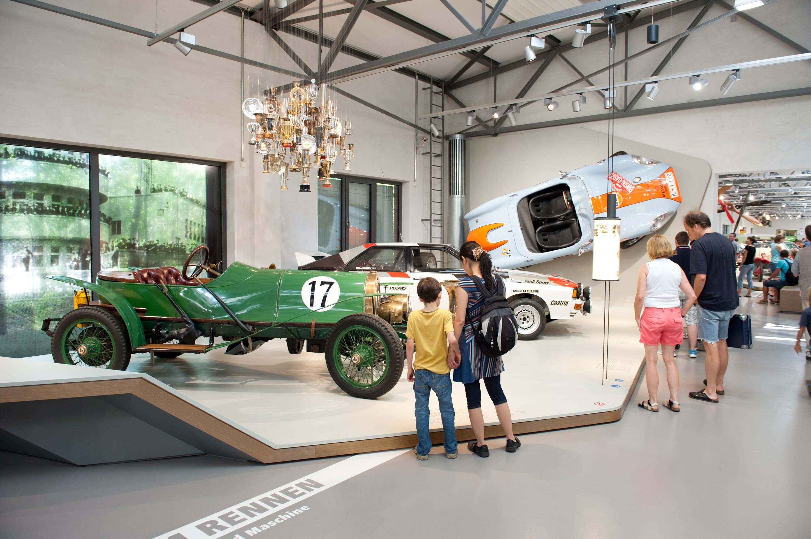 Blick in die Straßenverkehr-Ausstellung: Mehrere Personen stehen vor einem niedrigen Podest, auf dem zwei historische Rennwagen stehen. Ein weiterer blau-orangener Rennwagen ist an der Wand befestigt. Über dem Podest hängt eine Deckeninstallation aus zahlreichen Pokalen.
