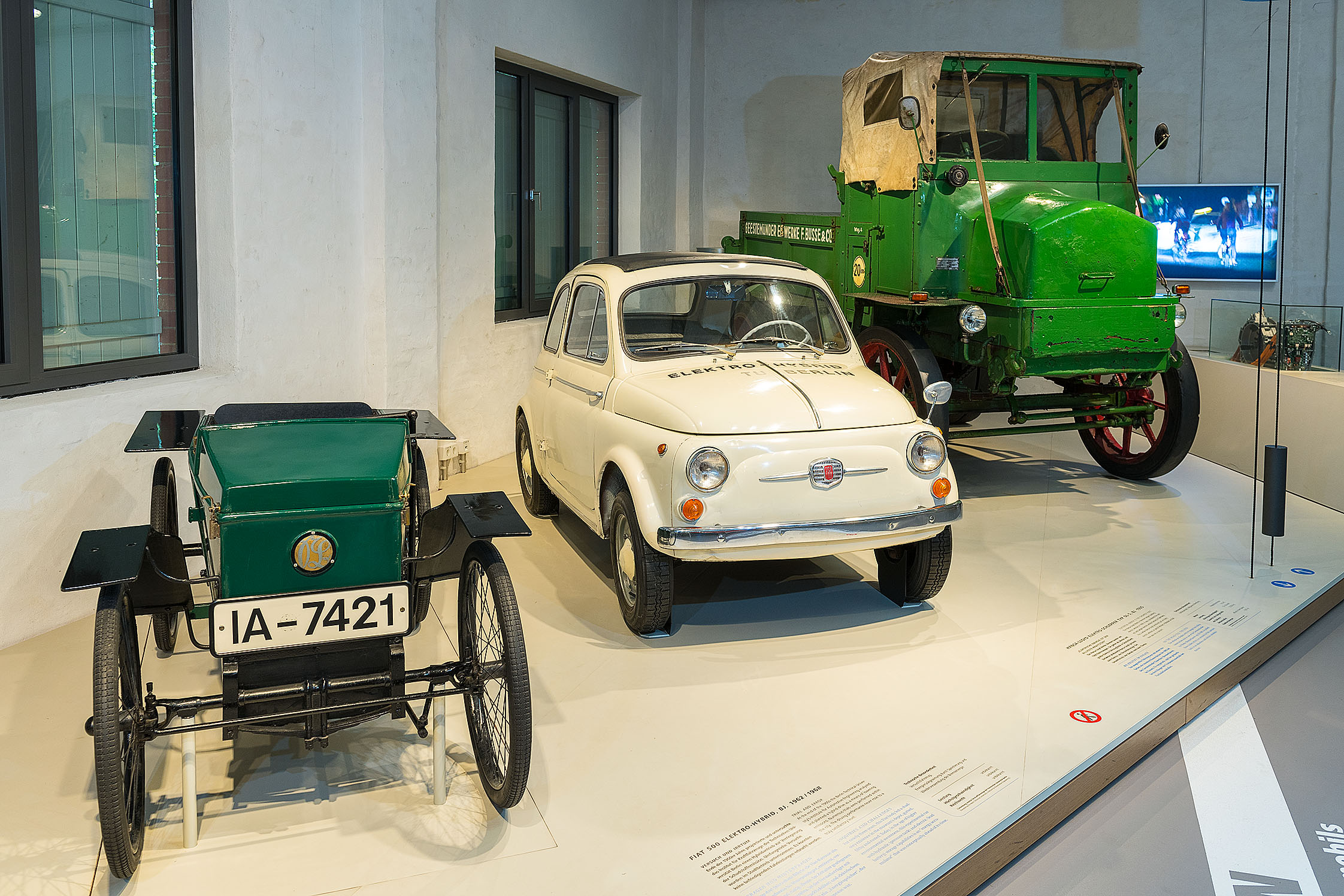Auf einem niedrigen Podest stehen drei historische Elektro-Fahrzeuge. Von links nach rechts: ein grüner offener Wagen, ähnlich einer Seifenkiste, ein weißer Fiat 500 und ein grüner LKW mit offener Ladefläche. 