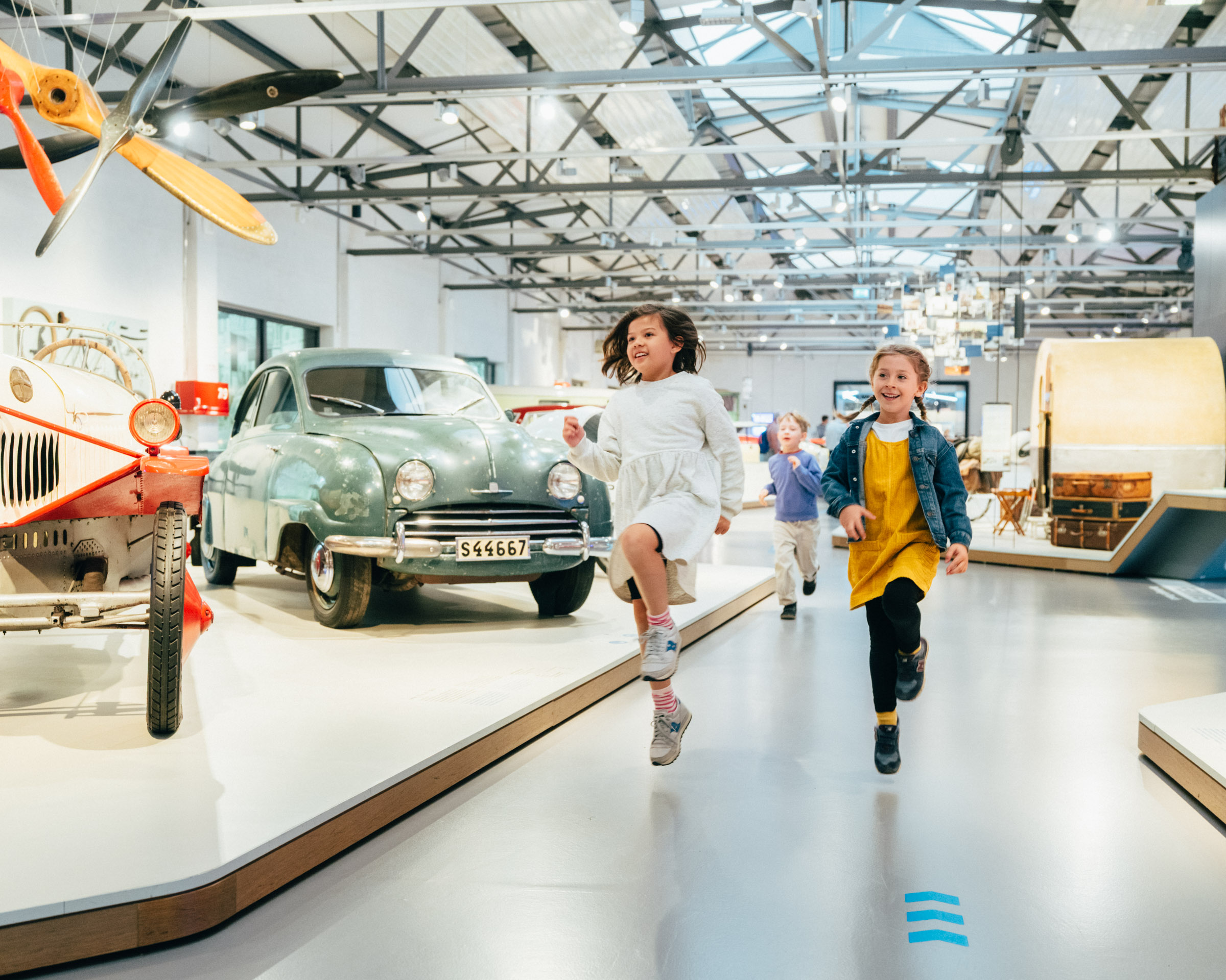 Blick in die Straßenverkehr-Ausstellung: Eine große Halle mit Stahlkonstruktion an der Decke. Ein breiter Gang zwischen niedrigen Podesten mit verschiedenen Fahrzeugen und Objekten.  Zwei kleine Mädchen laufen durch die Ausstellung.