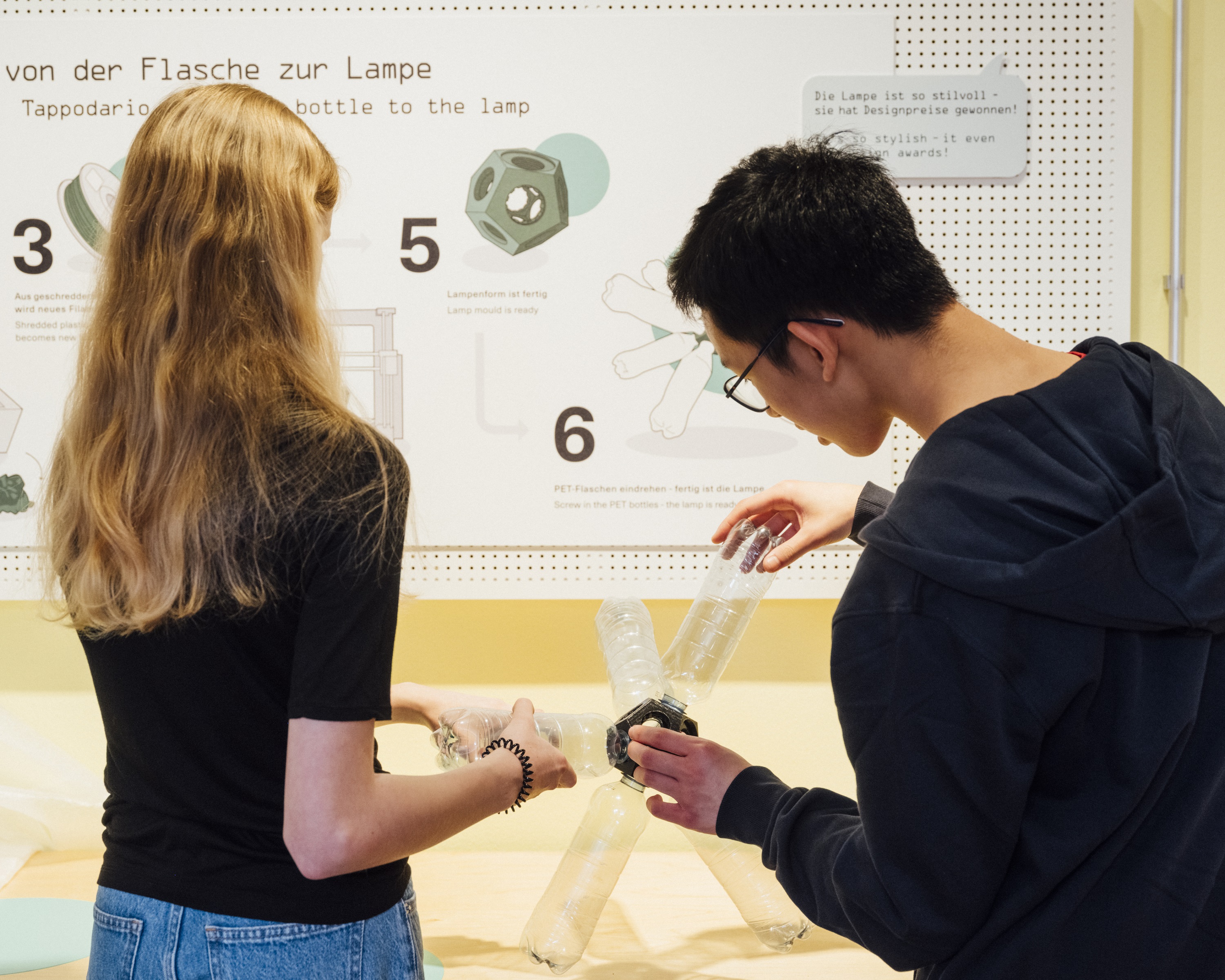 Vor einer Text- und Bildtafel mit der Aufschrift „von der Flasche zur Lampe“ schrauben ein Mädchen und ein Junge gemeinsam leere Plastikflaschen in eine Kunststoff-Fassung. Das Ergebnis ist eine lustig geformte Lampe.