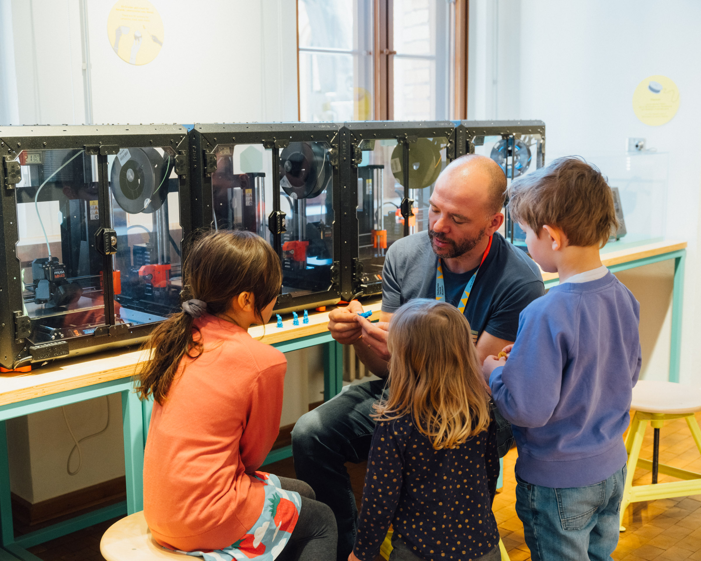 Ein Mann und drei Kinder sitzen vor einer Reihe von 3D-Druckern. Der Mann erklärt etwas, die Kinder haben Spaß und hören zu.