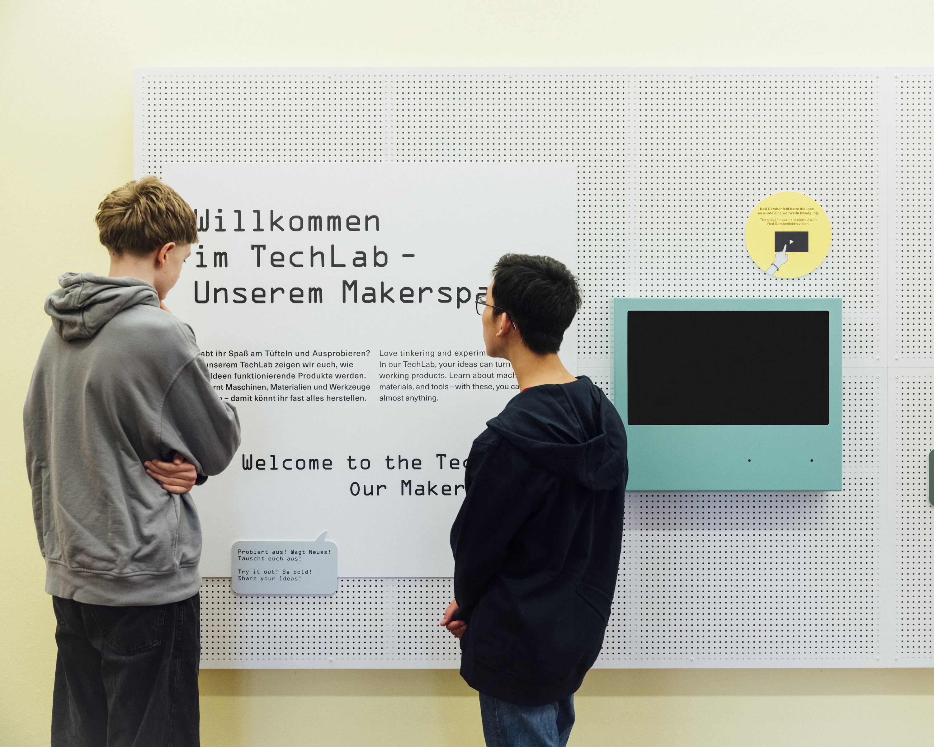 Zwei Jungen stehen vor einer Texttafel auf der die Überschrift „Willkommen im TechLab – unserem Makerspace“ auf Deutsch und Englisch zu erkennen ist. Sie lesen den Text. Daneben hängt ein Monitor mit blauem Rand.