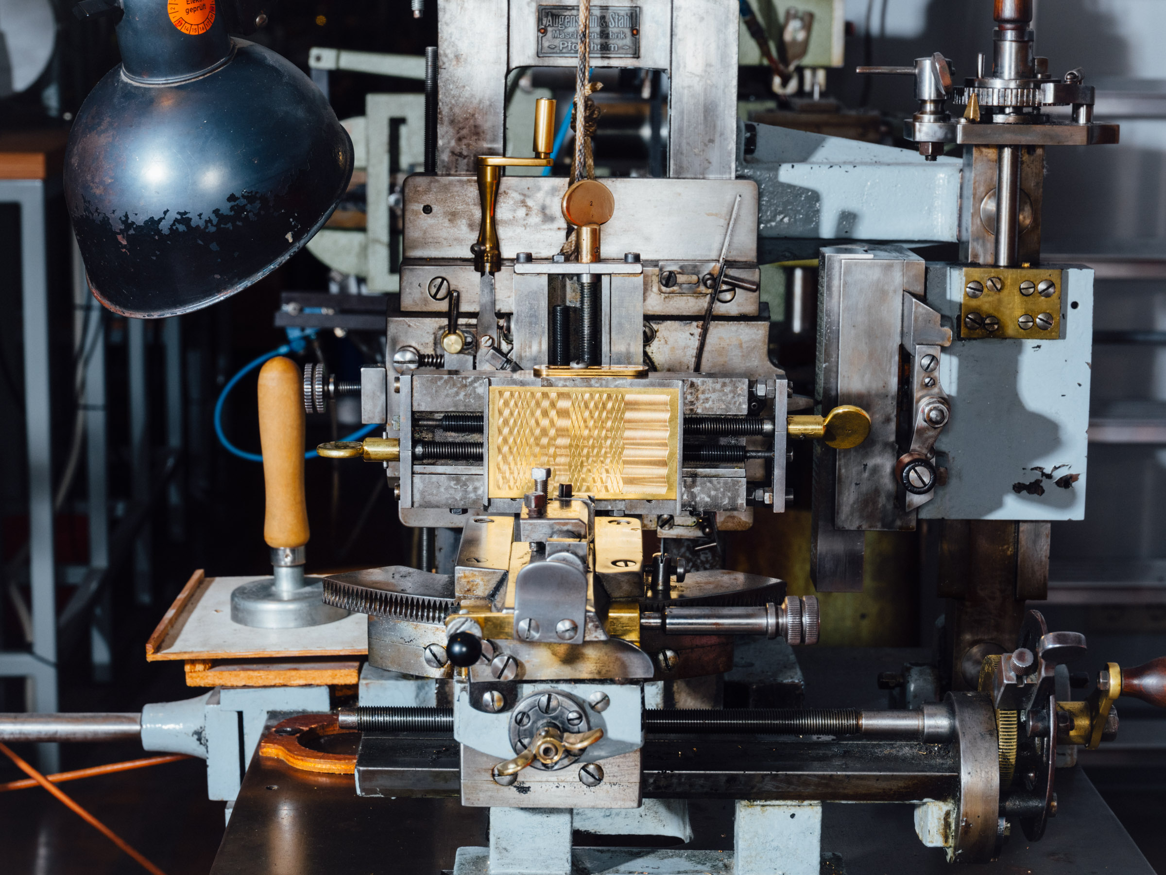 Eine Maschine aus Metall mit vielen Hebeln, an einer Stelle ist ein goldfarbenes Metallstück eingespannt, in das geometrische Muster eingeritzt sind.