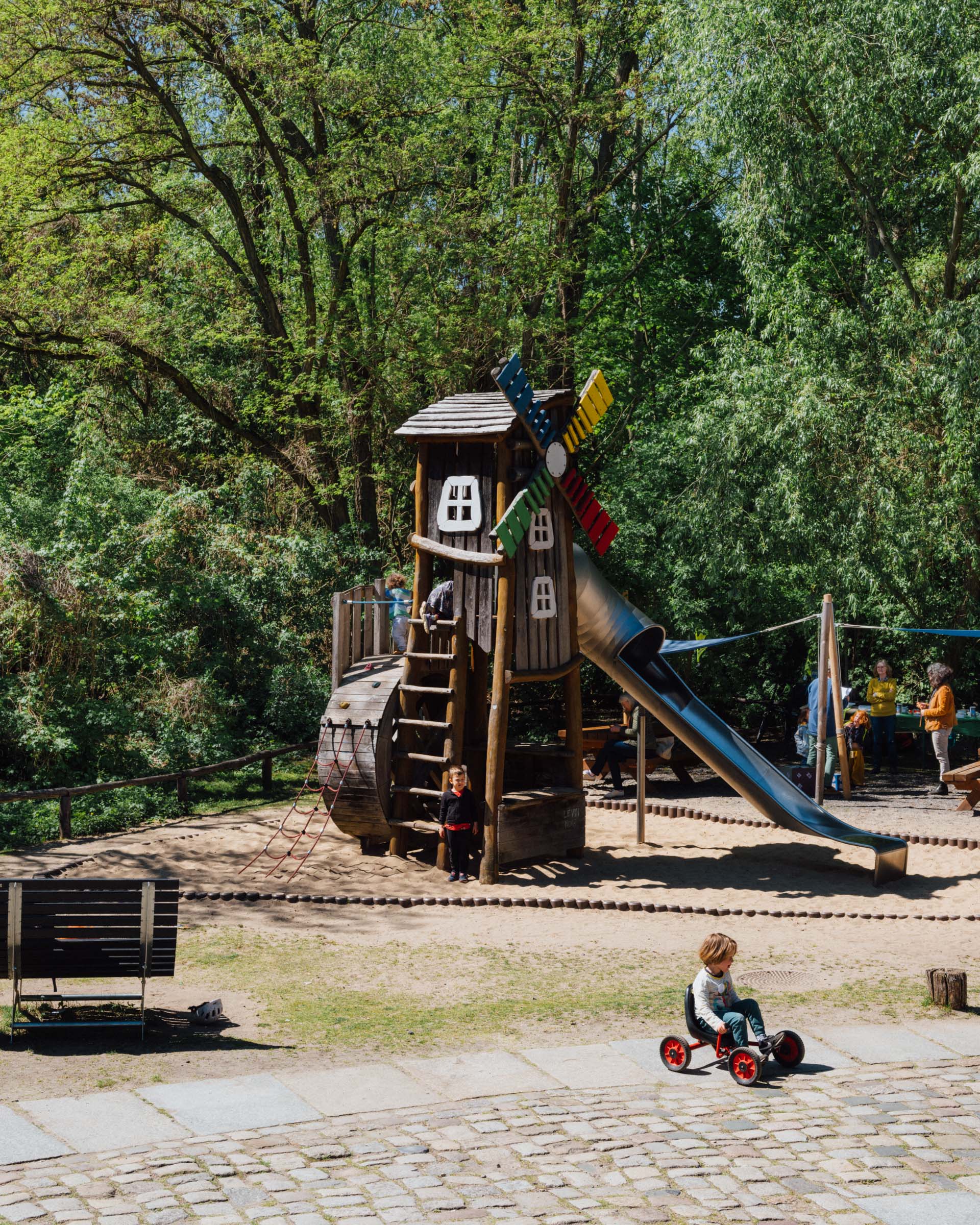 Ein Klettergerüst in Form einer Mühle steht auf einem Spielplatz mit Sandkasten. Darum herum Bäume. Auf dem Spielplatz spielen Kinder.