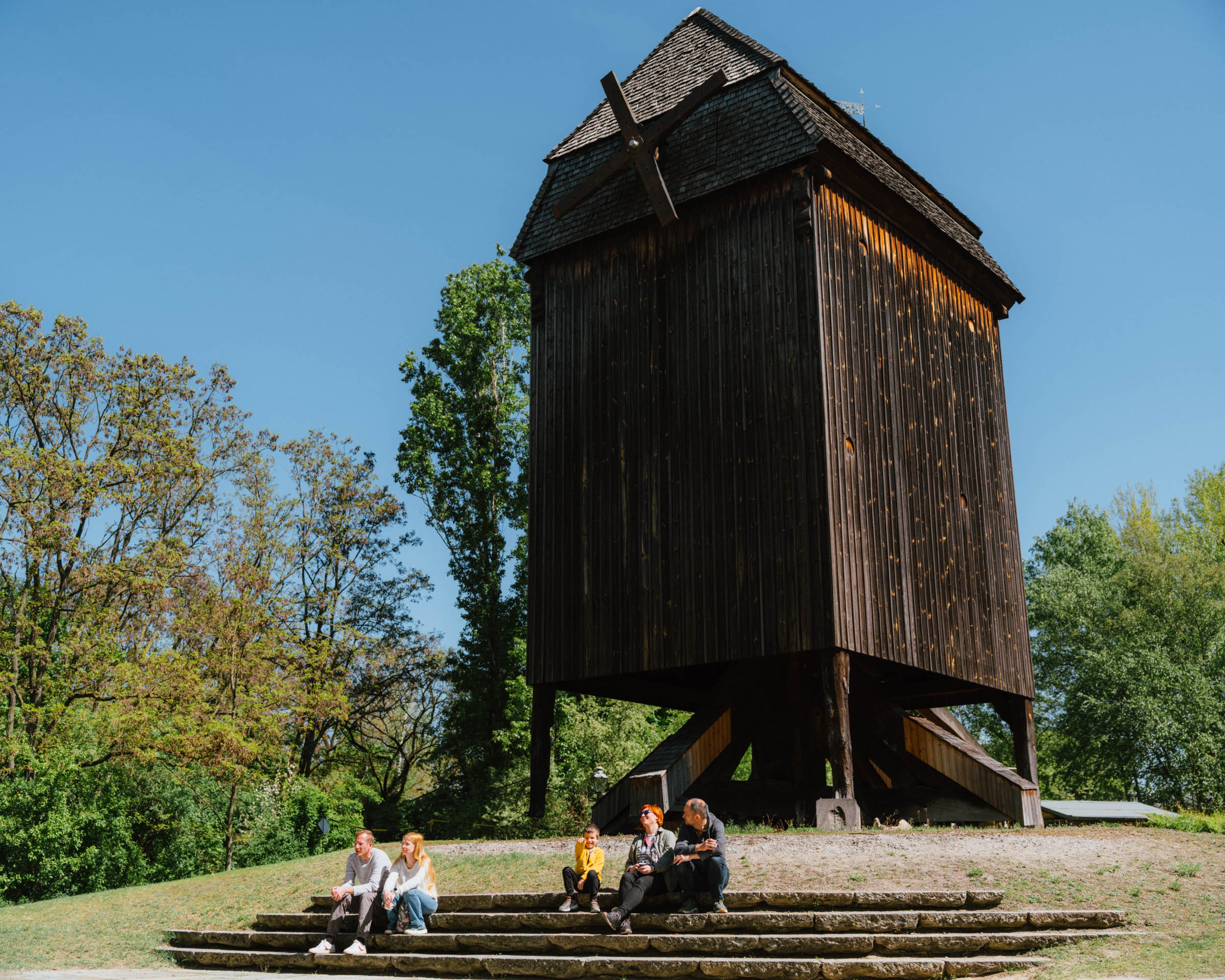 Eine Bockwindmühle in einem Park, auf den Stufen davor sitzen mehrere Menschen.