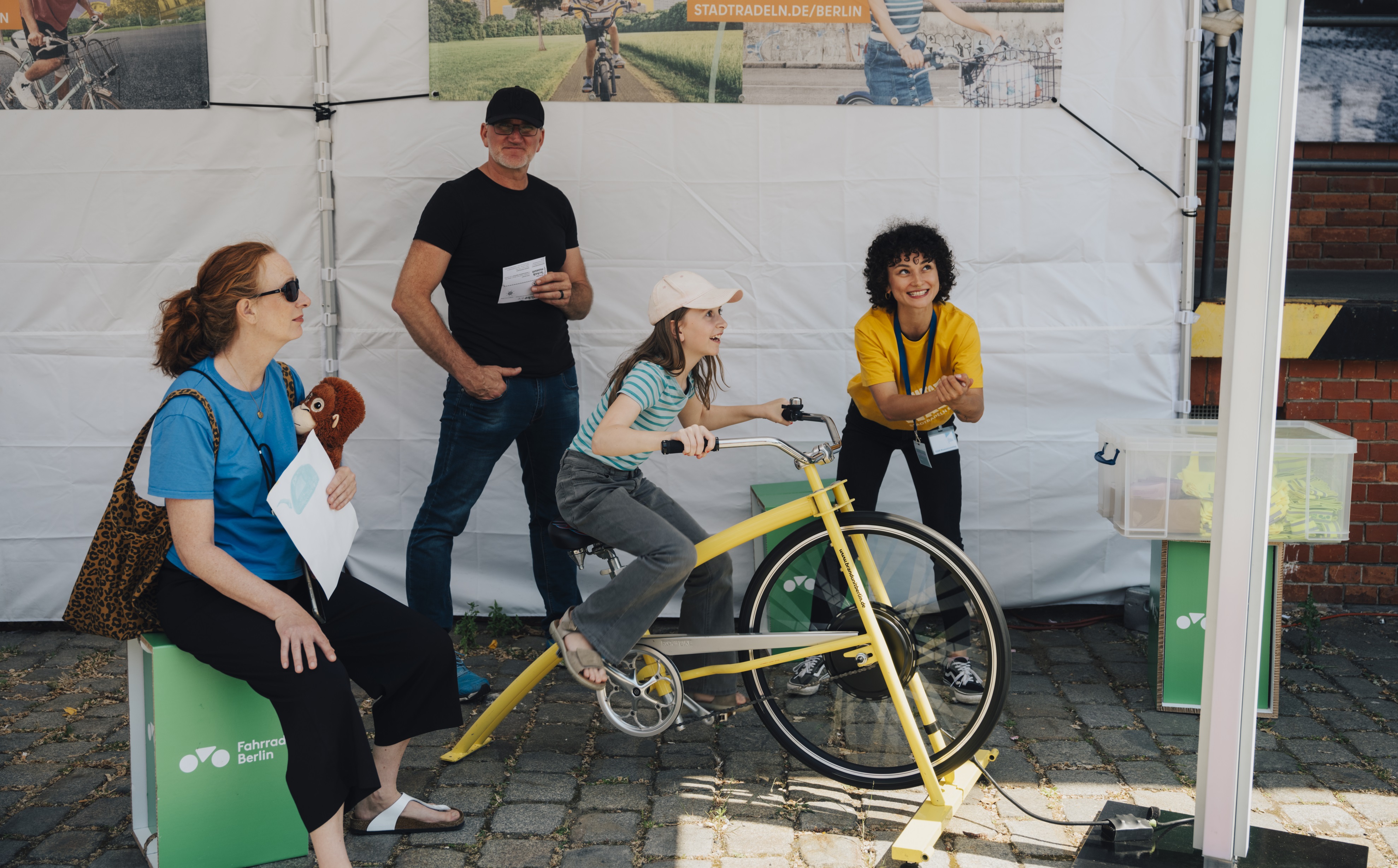 Mehrer Menschen stehen um ein fest montiertes Fahrrad herum, ein Mädchen fährt darauf.