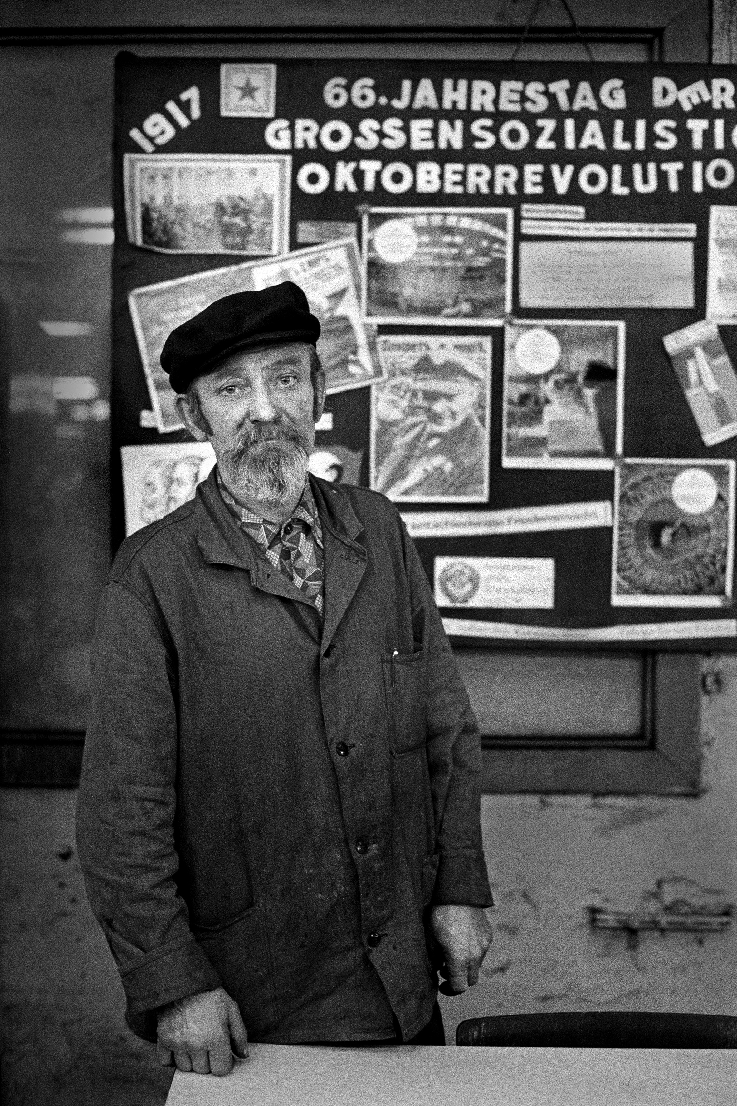Auf dem Foto ist ein Arbeiter mit Lenin-Mütze zu sehen, der vor einem schwarzen Brett mit kommunistischen Parolen und einem Foto von Lenin steht.