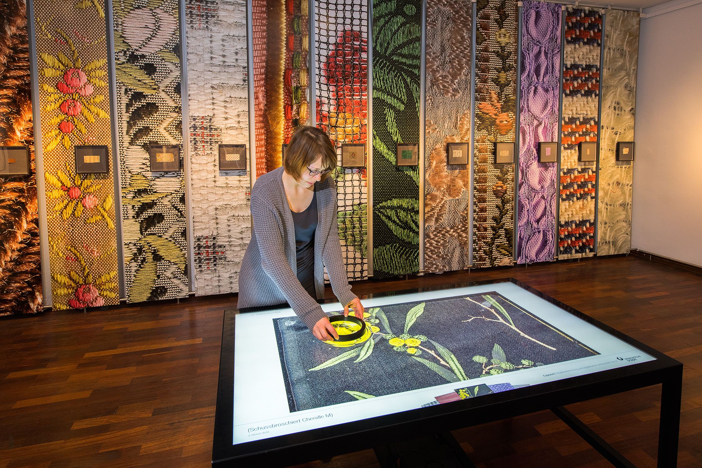 Blick in der Ausstellungsraum mit leuchtend bunten, hochauflösenden Detailaufnahmen von Stoffgeweben im extremen Hochformat. In der Mitte des Raumes steht eine Frau an einem Leuchttisch, auf dem man sich die Fotos der Stoffe stark heranzoomen kann.