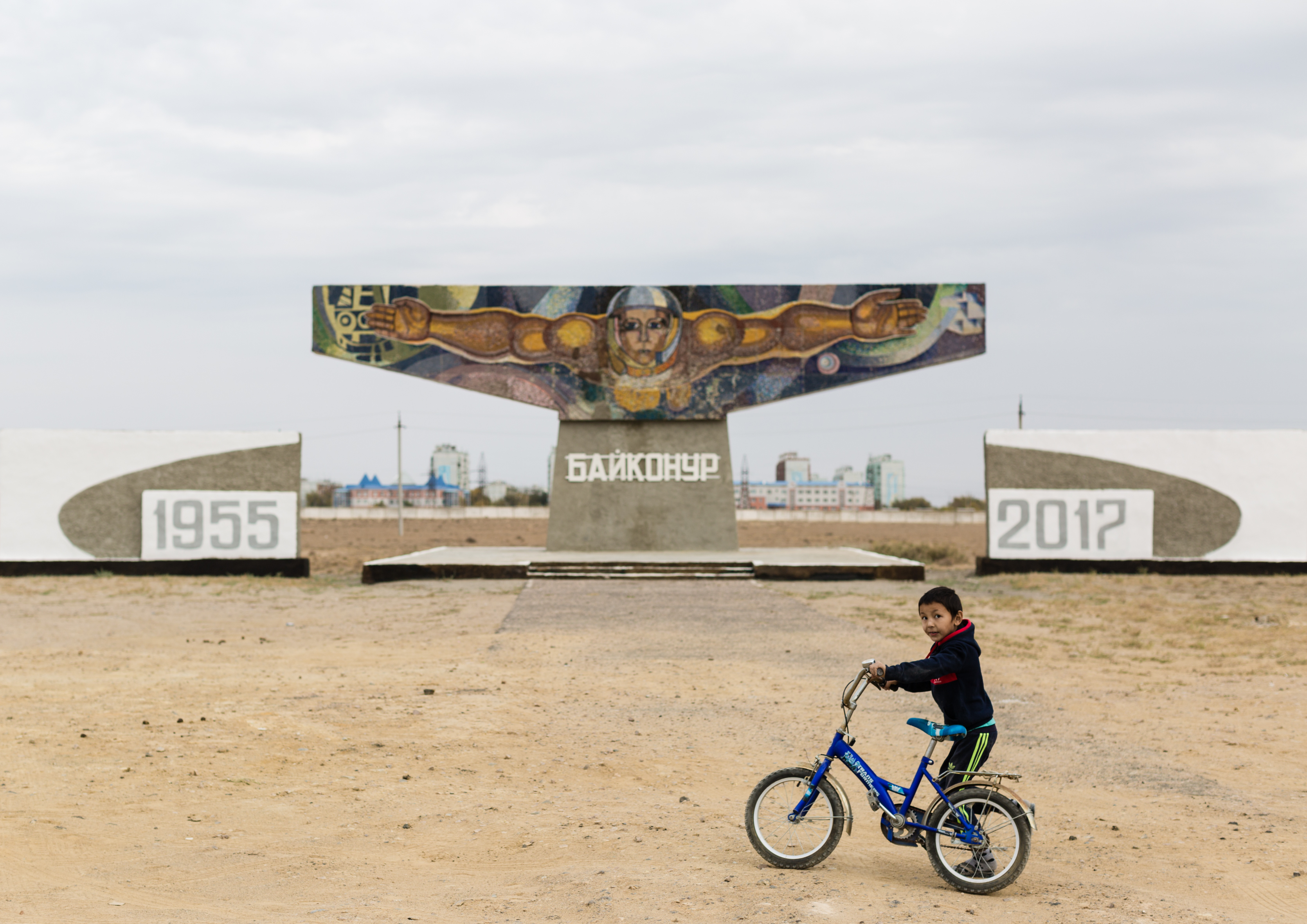 Ein kleiner Junge schiebt sein Fahrrad über einen großen sandigen Platz. Im Hintergrund steht ein großes Denkmal, darauf der Kopf einen Kosmonauten, der seine Arme weit ausbreitet.