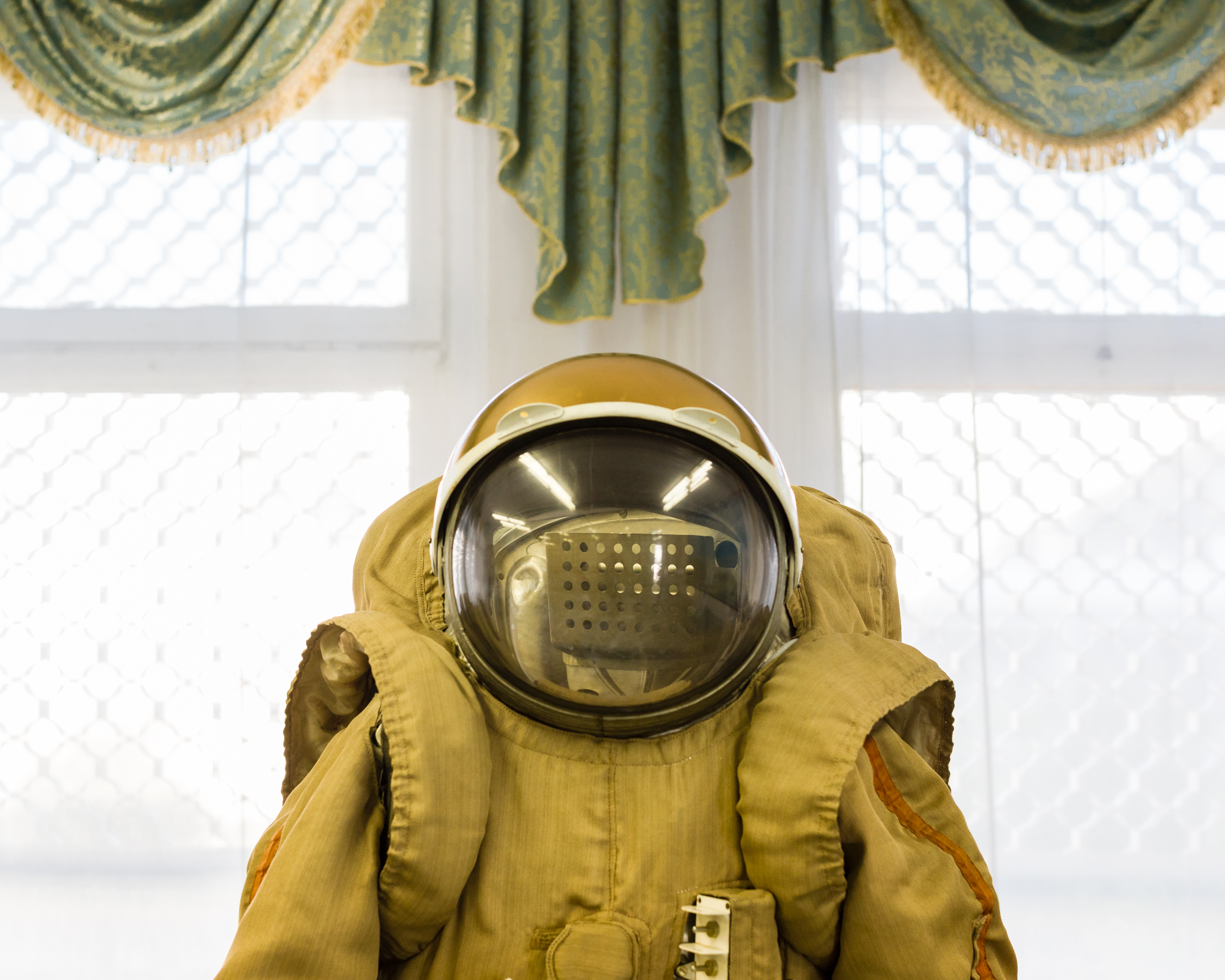Das Bild zeigt zentral einen Kosmonautenanzug von der Taille aufwärts. Darauf sitzt ein kugelförmiger Helm mit großem Glasvisier. Im Hintergrund hängt über einem Fenster eine Zierschabracke mit Goldbordüre.