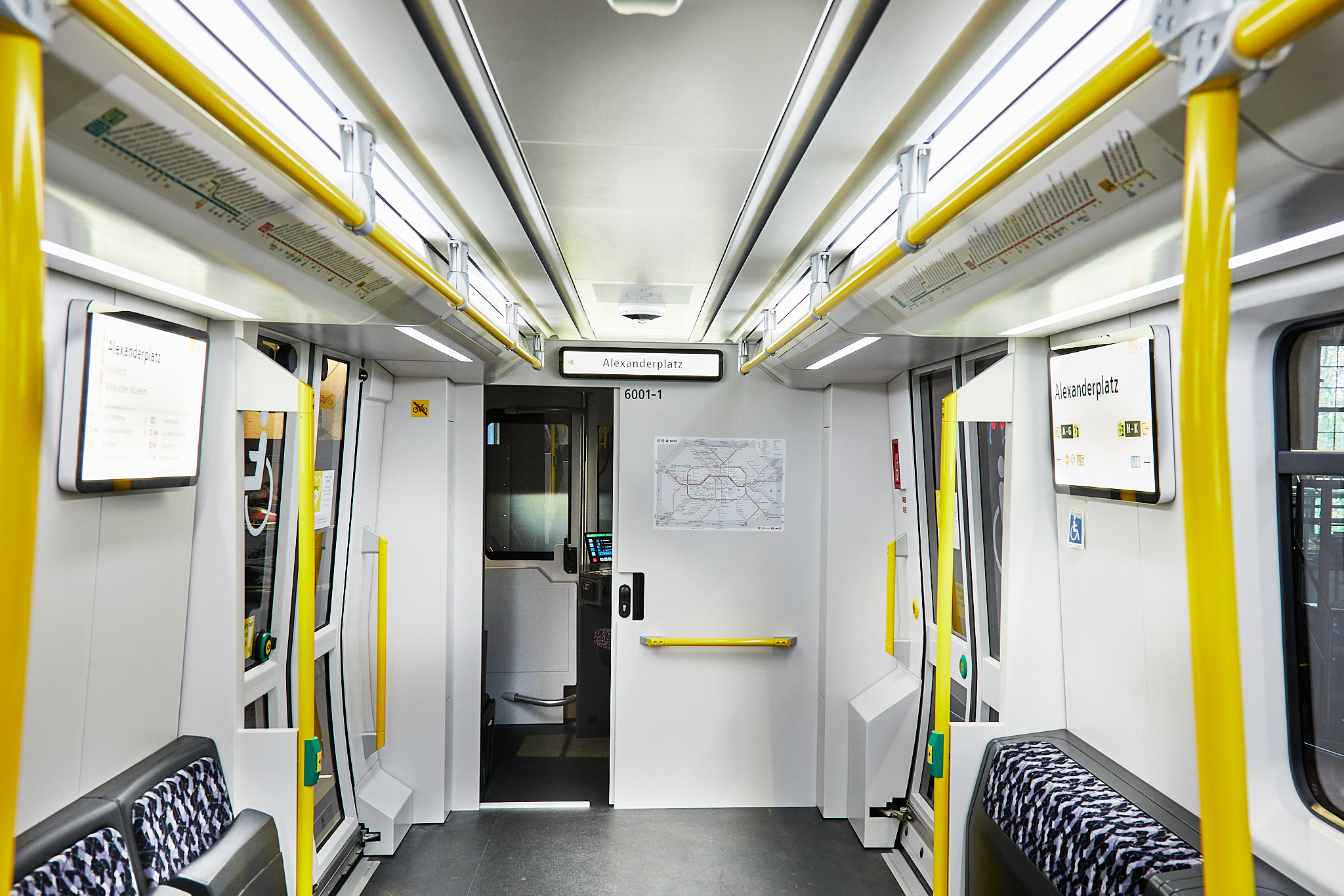 Blick in den Innenraum eines U-Bahnwagens. Der Eindruck ist hell und groß. Die Sitze sind blau-schwarz gemustert, die Haltestangen sind gelb.