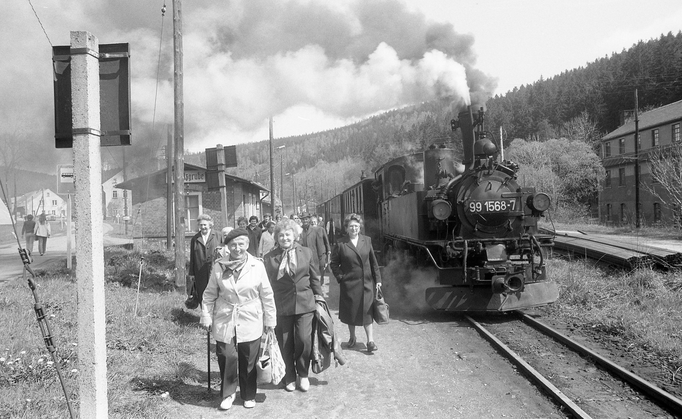 Ein ländlicher, kleiner Bahnhof mit einem kleinen, backsteinernen Bahnhofgebäude. Eine Gruppe von Menschen ist gerade mit einer dampfenden Lok angekommen und verlässt den Bahnhof. Einige ältere Damen lächeln dabei heiter.
