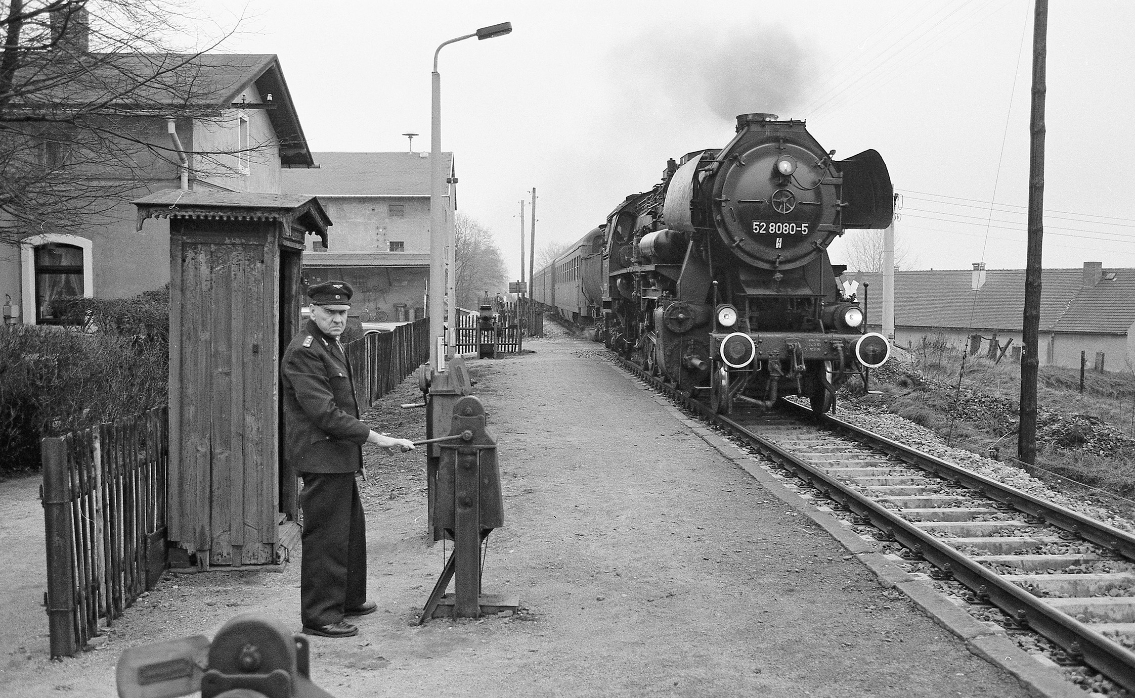 Eine Schwarzweiß-Fotografie: Auf einem Bahnsteig in ländlicher Umgebung steht ein älterer Schrankenkwärter in Uniform. Von hinten nähert sich eine Dampflokomotive.