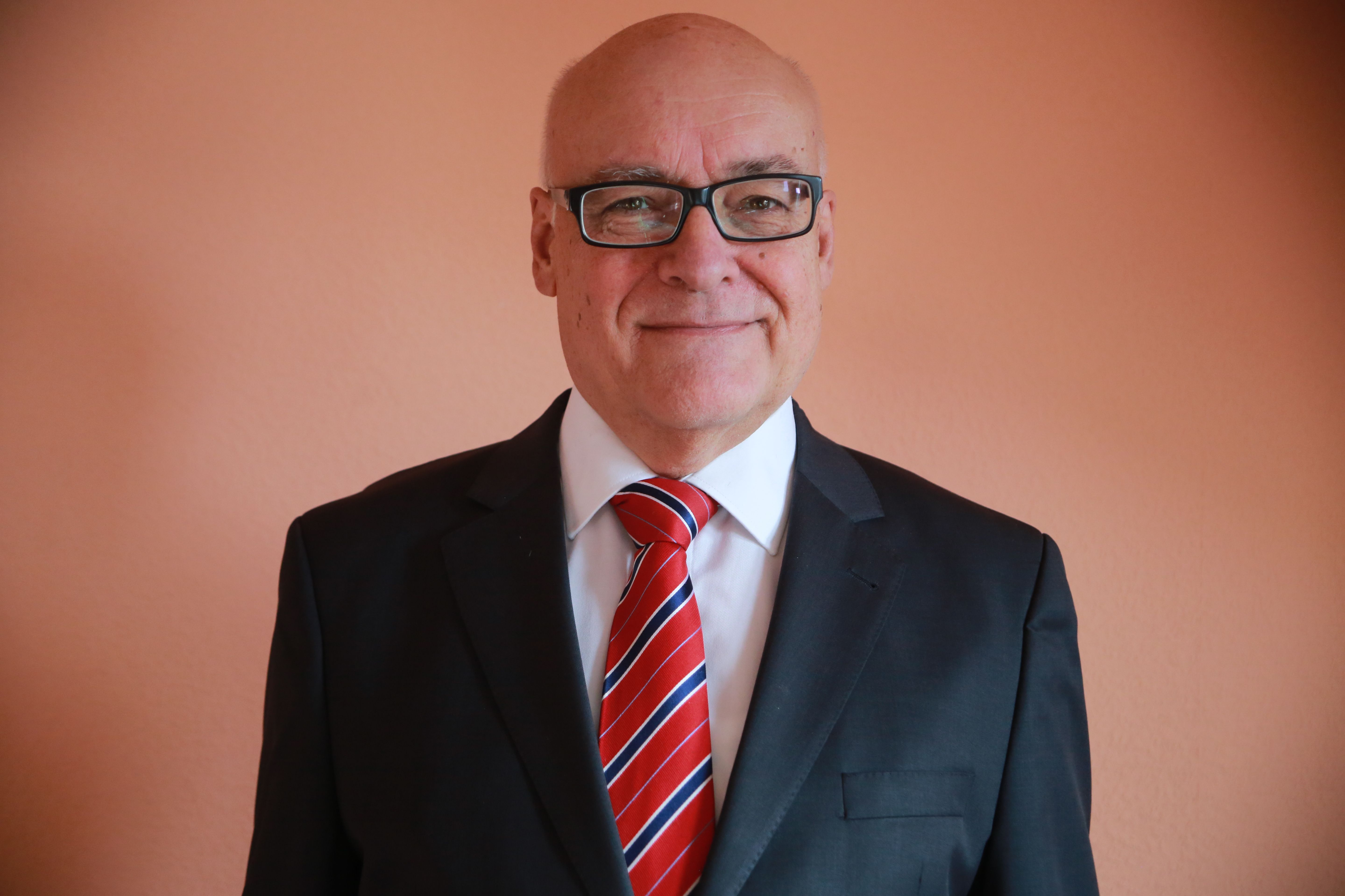 Das Bild zeigt eine Portraitaufnahme des Fotografen Burkhard Wollny, wie er im schwarzen Anzug vor einer rötlichen Wand steht.