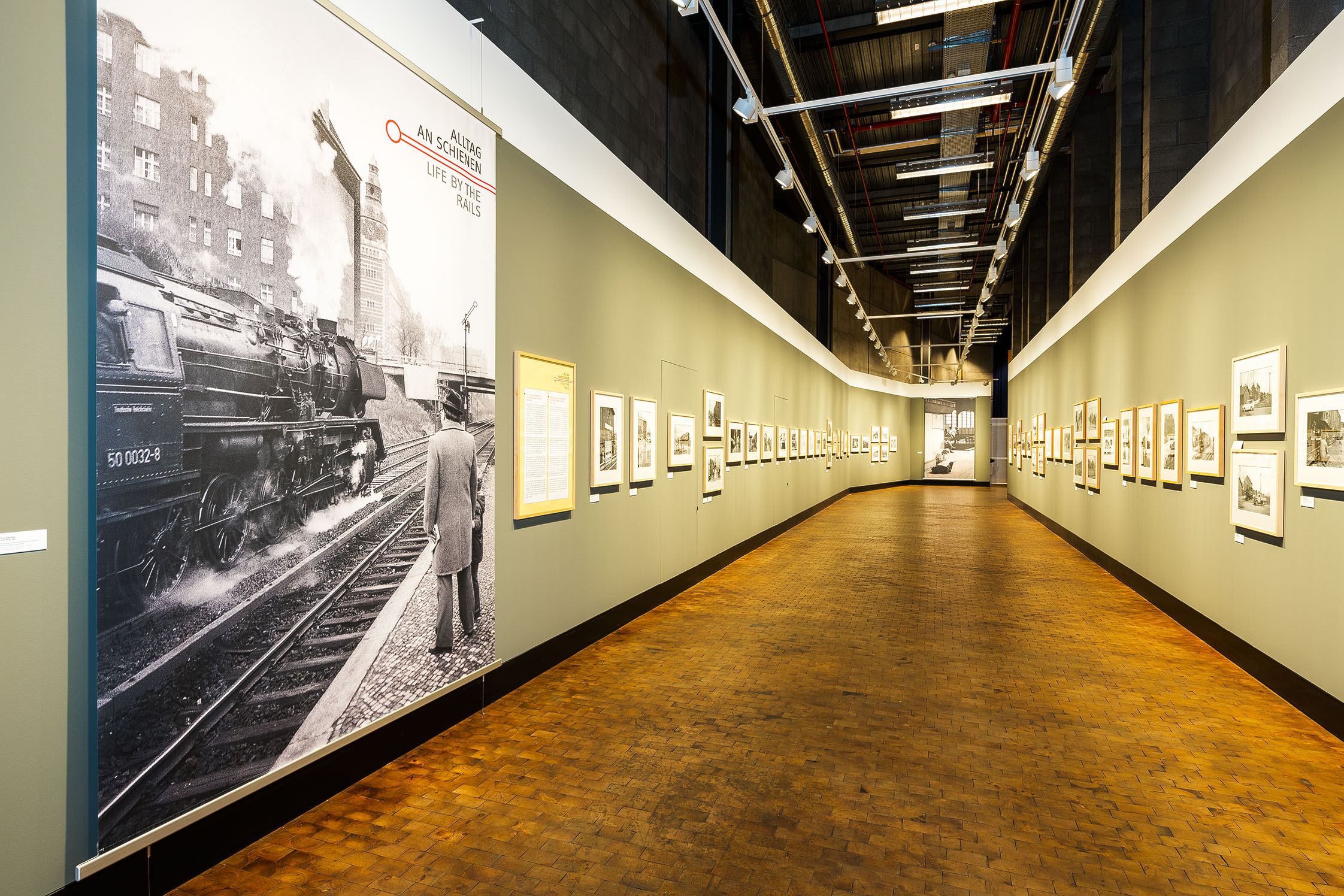 An beiden Seiten des langen Gangs der großen Galerie hängen Schwarz-Weiß-Fotos an den grau-grünen Wänden. 