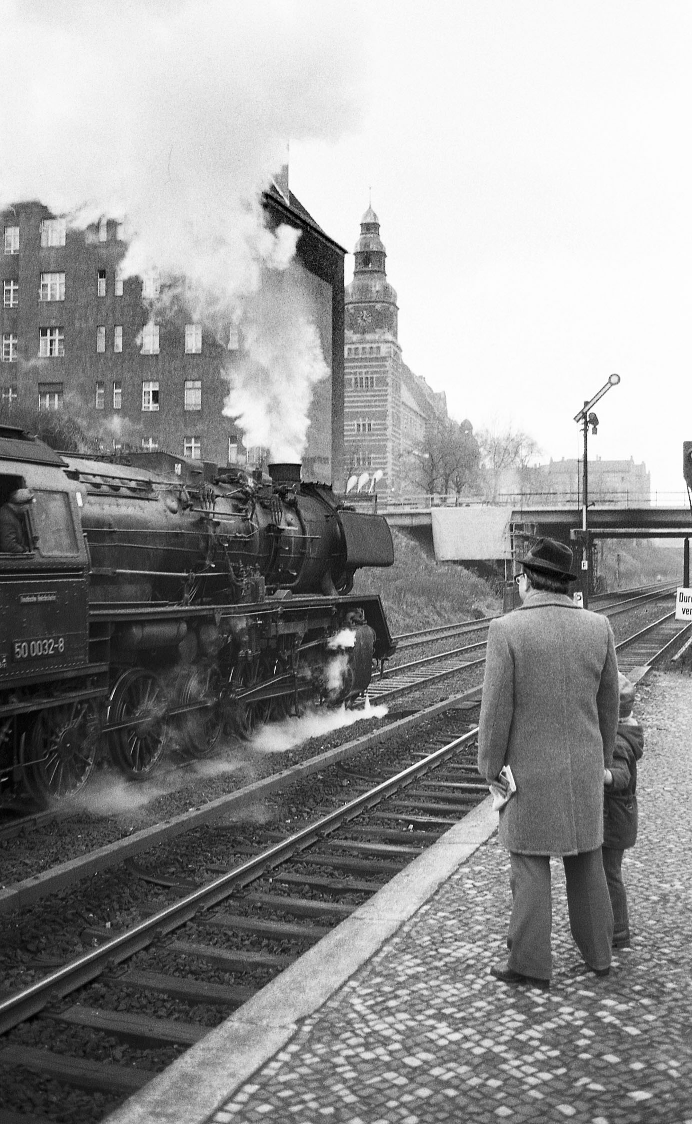 Ein Mann und ein Kind stehen auf einem Bahnsteig und schauen der dampfenden Reko-Lokomotive nach, die an ihnen vorbei rauscht.