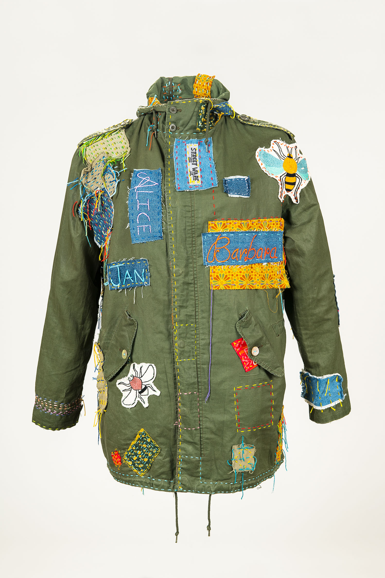 Eine olivgrüne Outdoorjacke ist mit diversen auffälligen farbenfrohen Patches repariert: Ins Auge stechen zwei Bügelflicken mit Insektenmotiven, außerdem großflächig gestickte Namen auf Bügelflicken, die auf die Beteiligten des Berliner Kollektivs "STREETWARE saved item" hinweisen.  