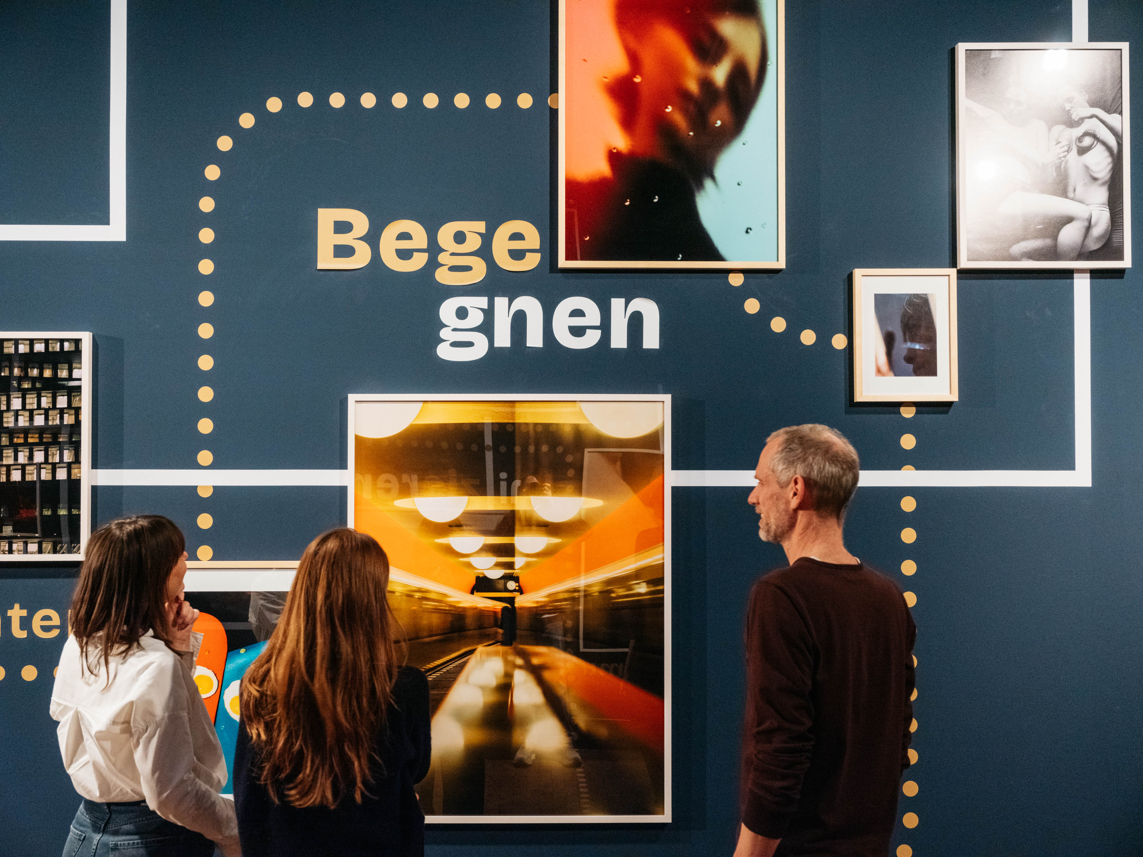 An einer dunkelblauen Wand hängen mehrere gerahmte Bilder. In der Mitte ist das Wort "Begegnen" zu lesen. Zwischen den einzelnen Fotografien sind gepunktete Verbindungen und Verbindungslinien zu sehen. Drei Personen stehen vor den Bildern und betrachten diese.