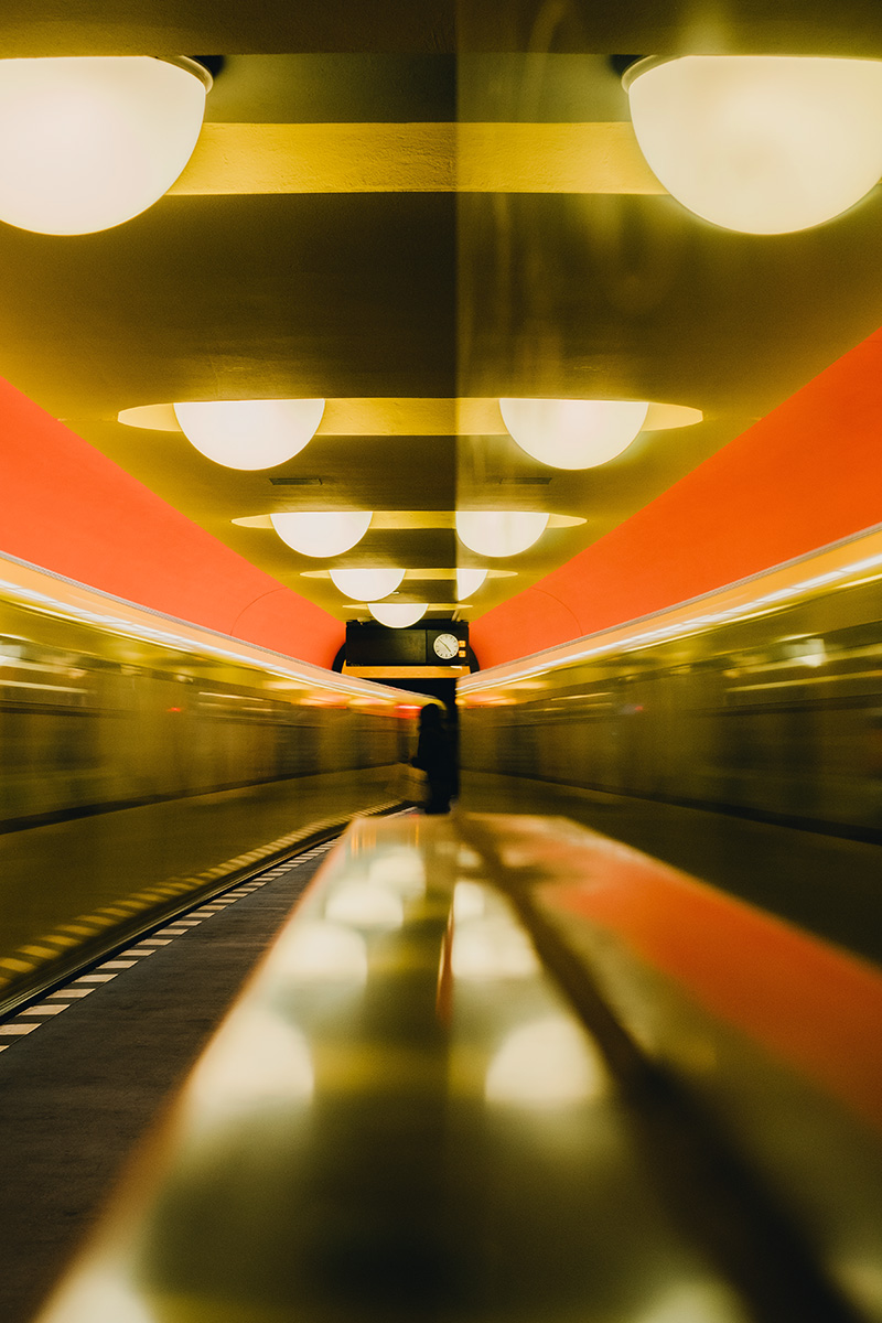 Das Farbfoto zeigt eine U-Bahn-Station mit halbkugelförmigen Deckenleuchten. Auf der linken Seite fährt gerade ein Zug durch. Auf dem zentralen Bahnsteig erkennt man die Silhouette eines Menschen, der unter der Anzeigetafel mit Uhr steht. Die rechte Bildseite wir von einer Fläche eingenommen, auf welcher sich die die Szenerie verschwommen widerspiegelt.