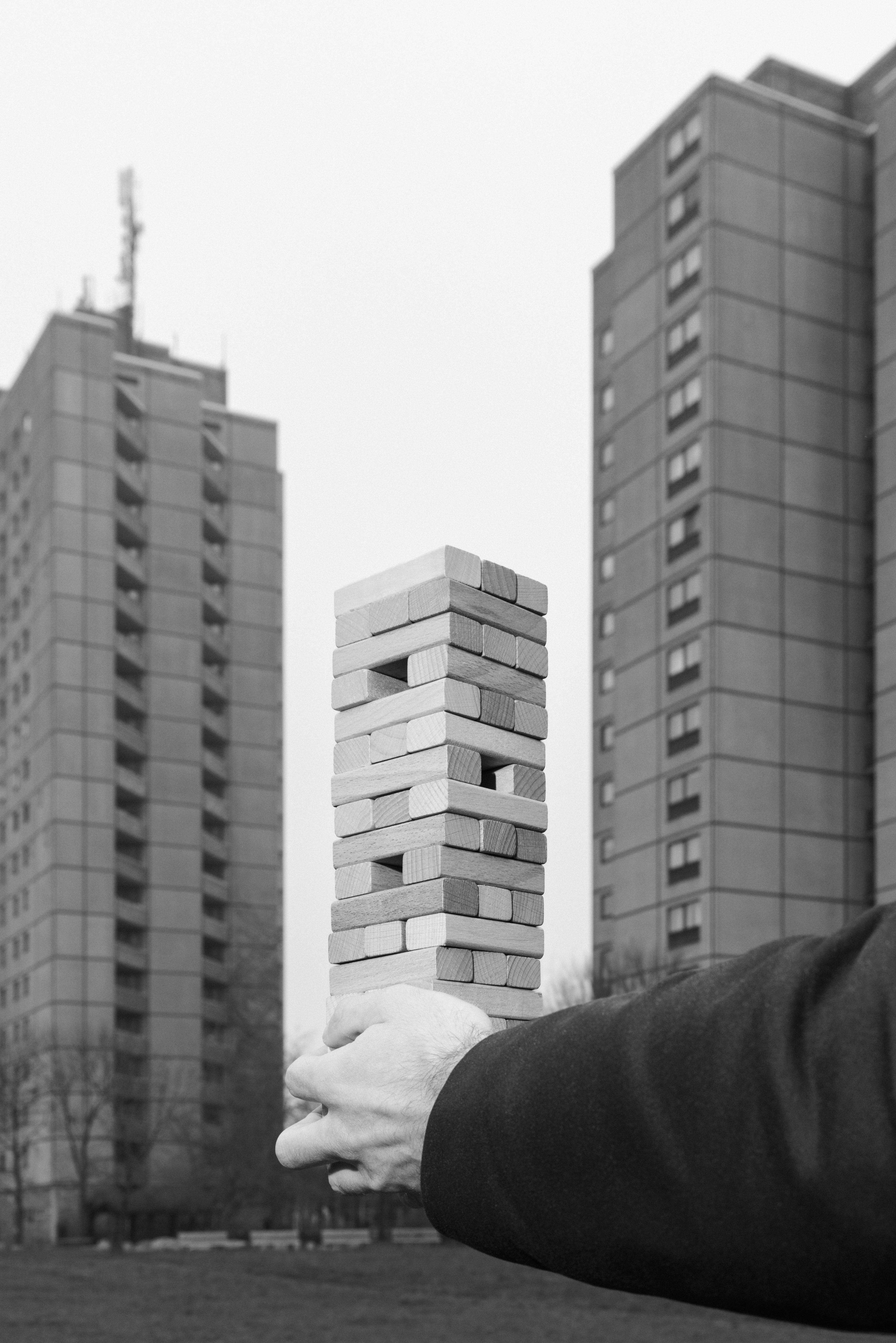 Das schwarz-weiße Foto zeigt eine Hand, die einen Jenga-Turm aus Holzklötzen hält. Im Hintergrund stehen zwei Hochhäuser, die optisch mit dem Turm korrespondieren. Durch die Perspektive wirkt es, als wäre der Jenga-Turm Teil der Architektur.