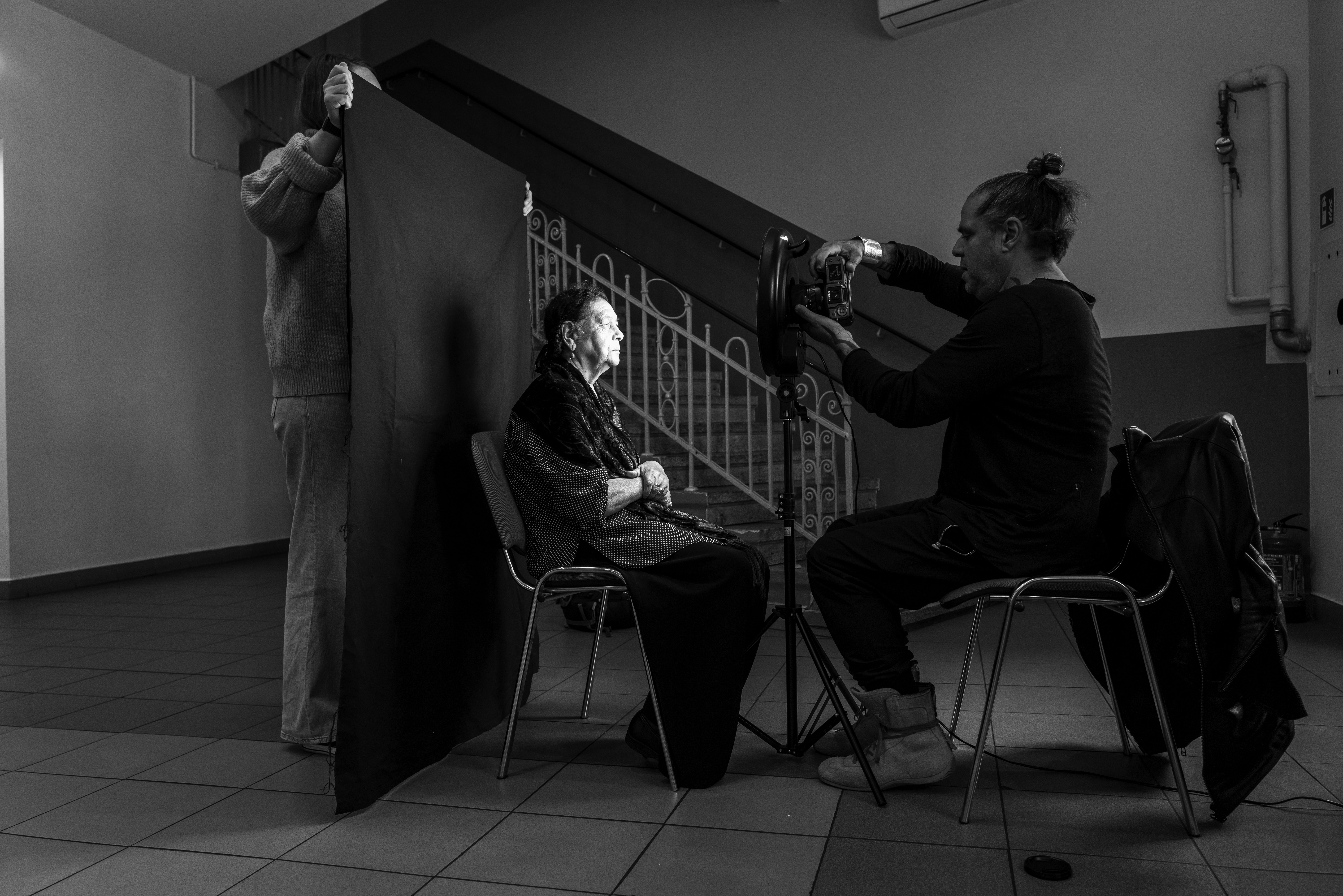 Schwarz-Weiß-Fotografie. Eine Person sitzt auf einem Stuhl. Hinter ihr hält eine weitere Person eine schwarze Leinwand hoch. Vor ihr sitzt eine weitere Person auf einem Stuhl und fotografiert die erste Person.