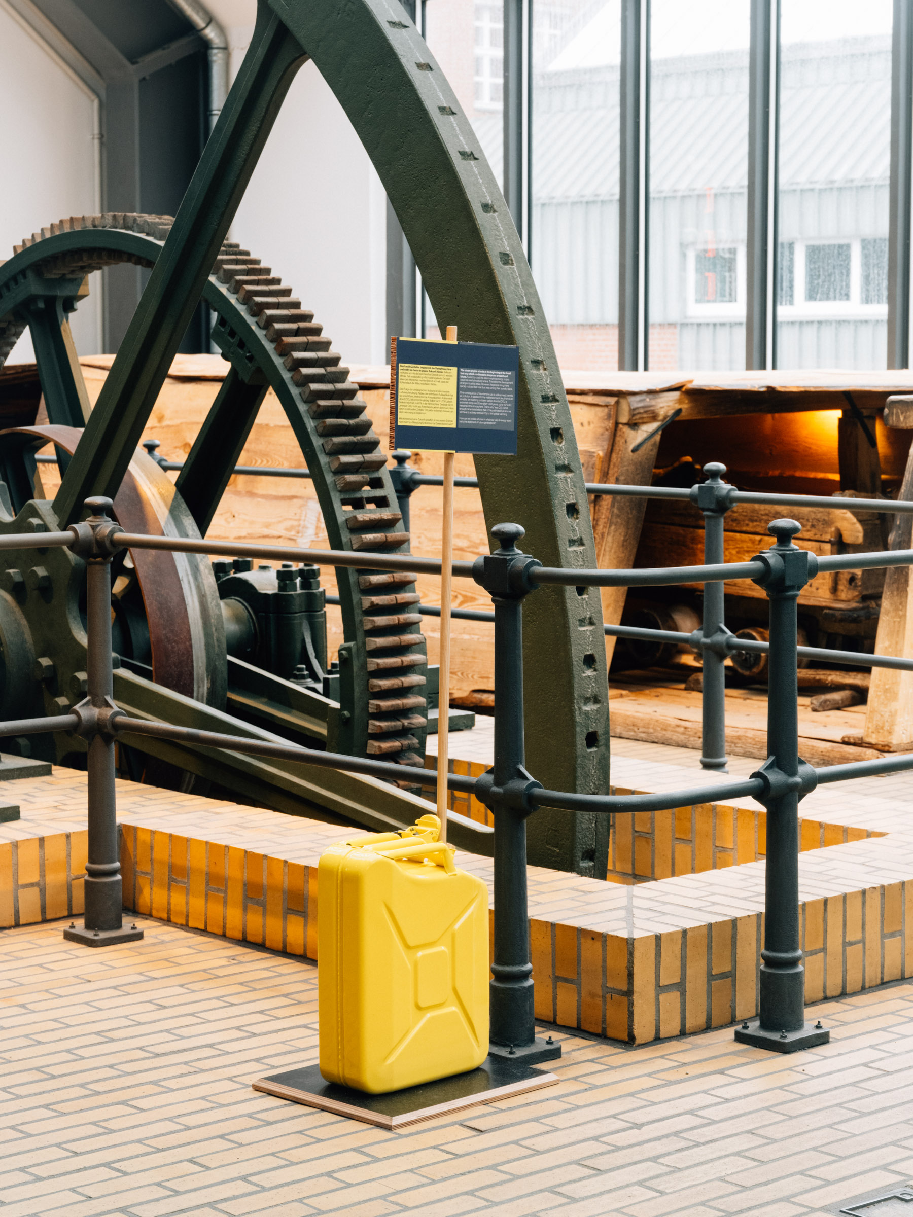 Vor einer Balancier-Dampfmaschine im Museum steht ein gelber Kanisterter und ein Objektschild.