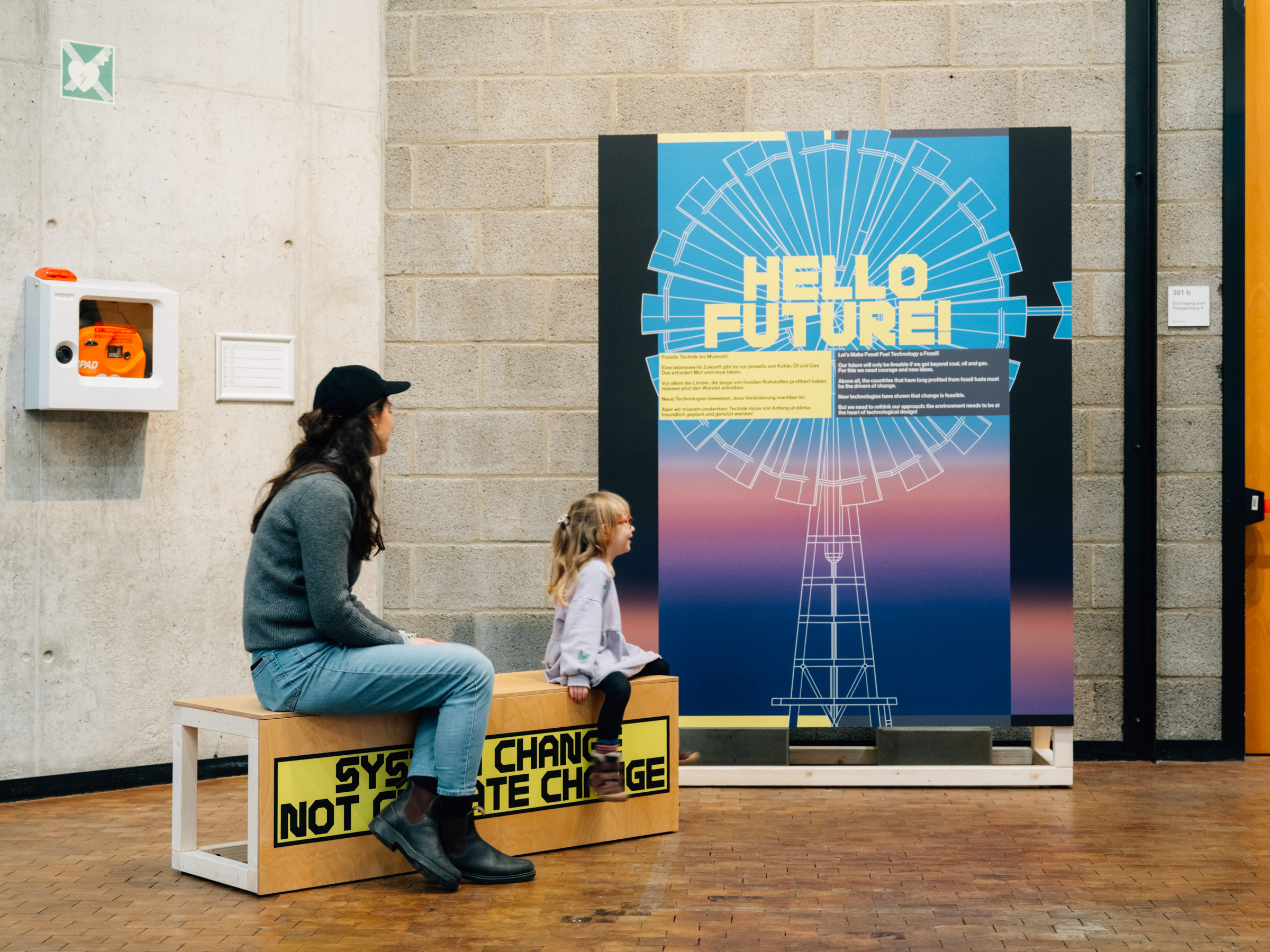 Eine Frau und ein kleines Mädchen sitzen auf einer Bank, die mit dem Slogan "System change, not climate change" beschriftet ist. Auf einem Plakat steht "Hello Future".