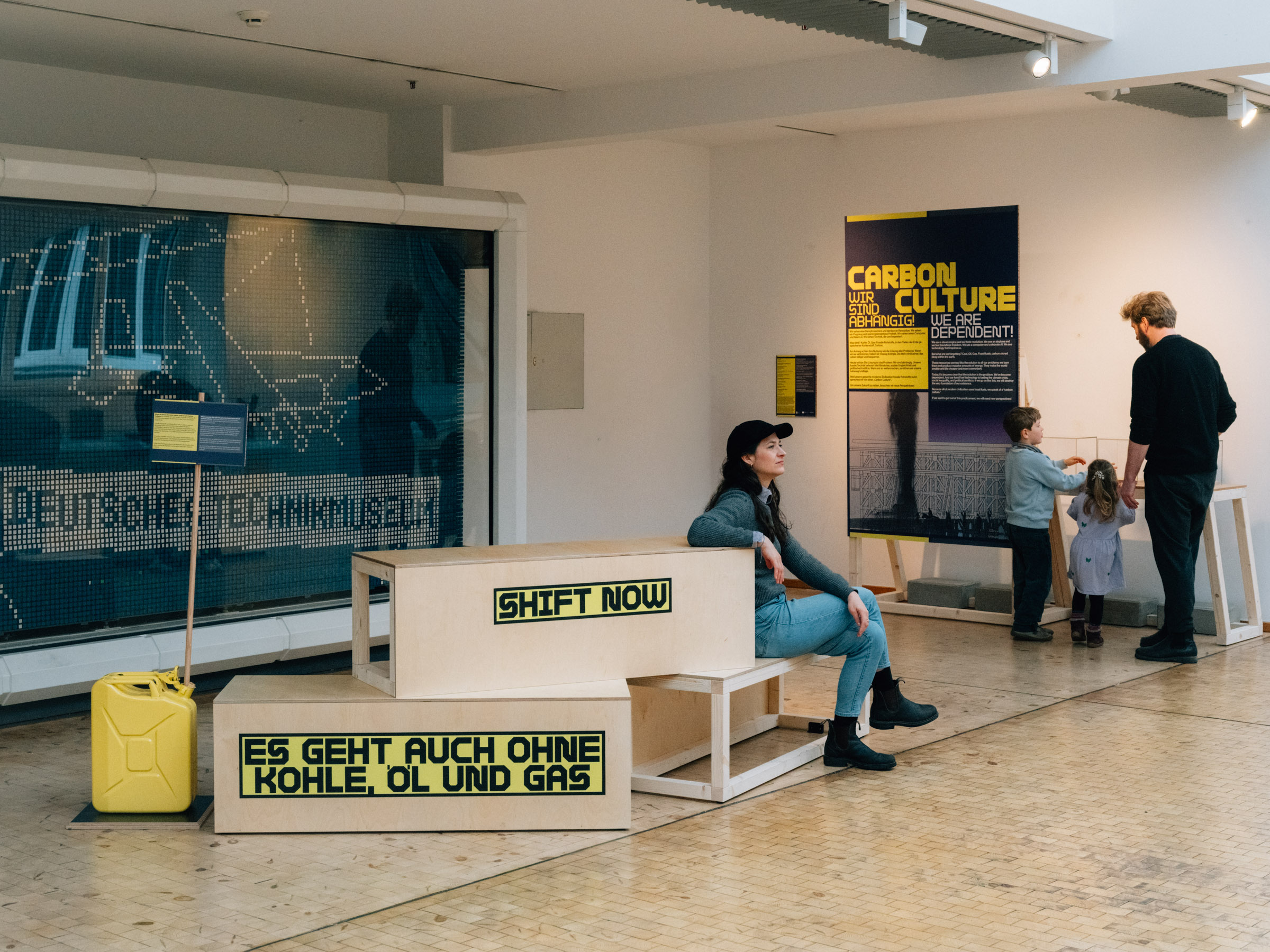 Mehrere Menschen halten sich in einem Ausstellungsbereich auf. Auf Sitzmöbeln steht "Shift now" und "Es geht auch ohne Kohle, Öl und Gas". Auf einem Plakat steht "Carbon Culture". Außerdem ist ein gelber Kanister zu sehen.