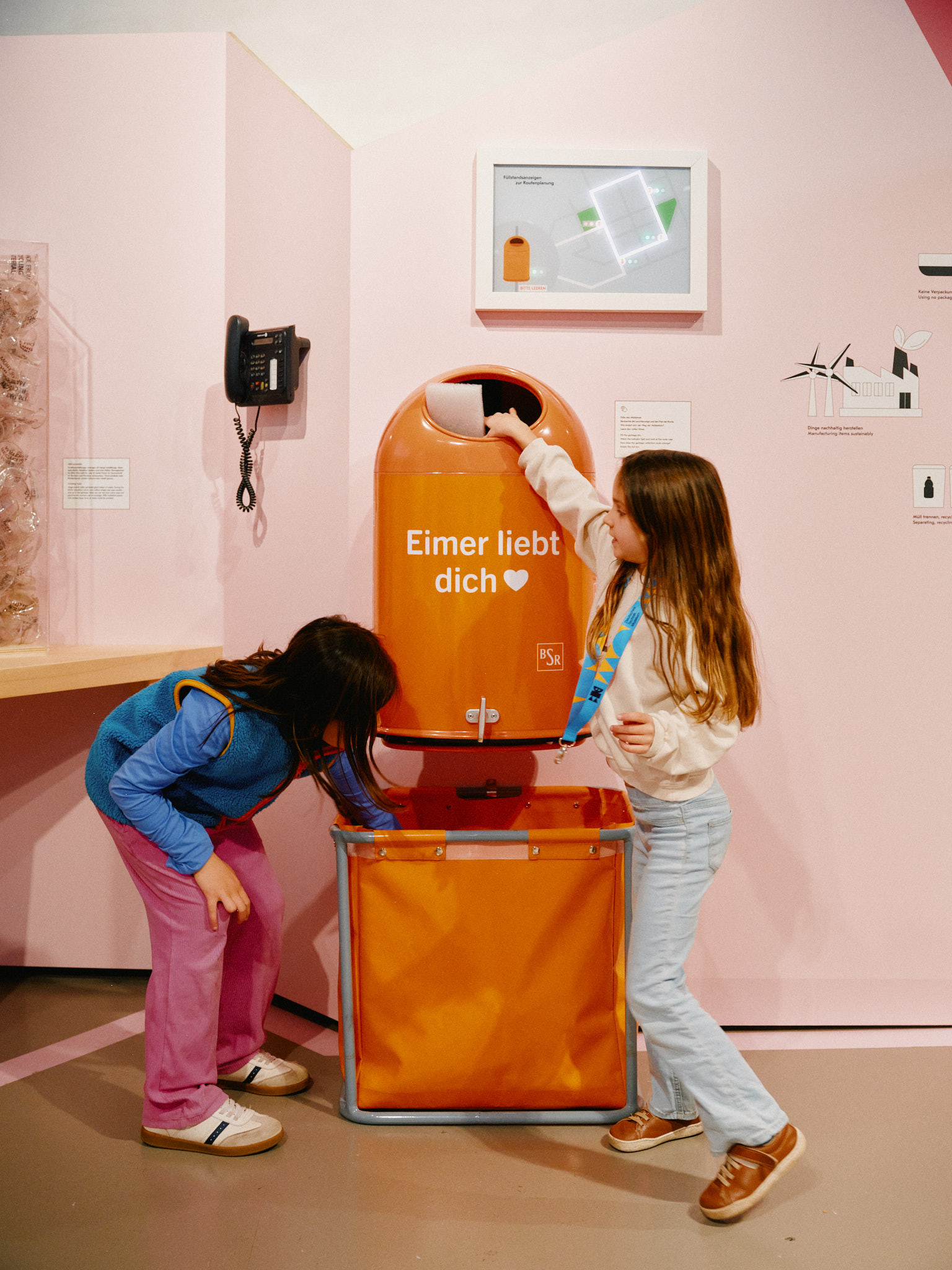 Ein orangener Mülleimer, auf dem „Eimer liebt Dich“ steht. Zwei Mädchen befüllen den Eimer, darunter eine orangefarbene Tasche, die den Müll beim Entleeren auffängt. Dahinter ein Display, das zeigt, wann der Mülleimer voll ist.