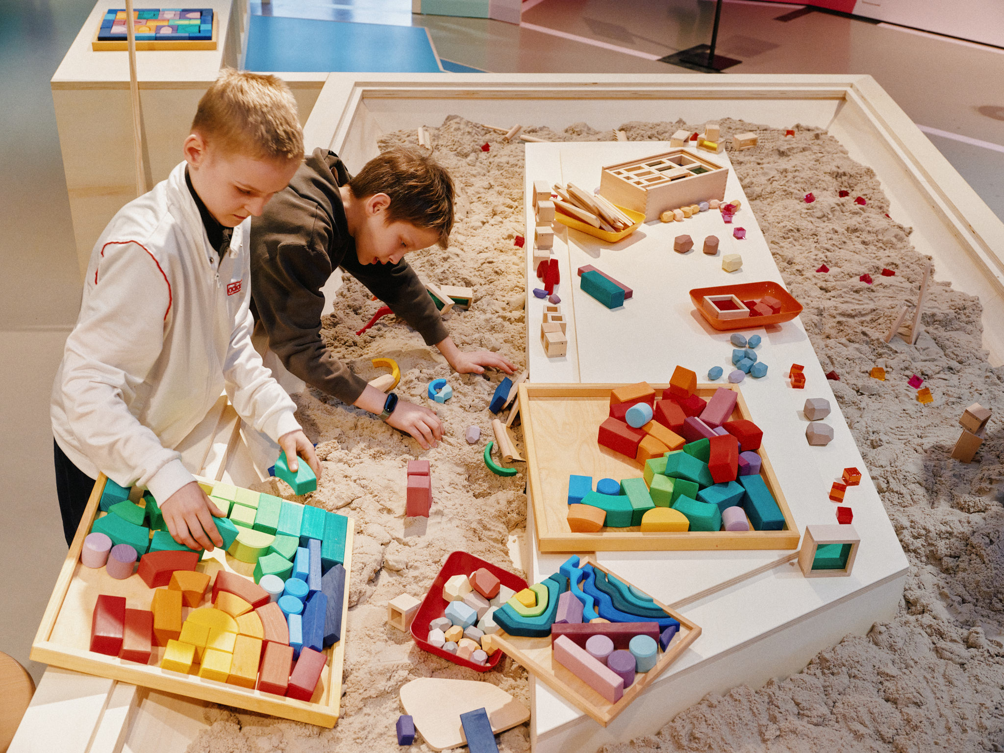 In einem großen Sandkasten liegen viele bunte Bauklötze. Zwei Jungen bauen mit dem Sand und den Bauelementen eine kleine Stadt.