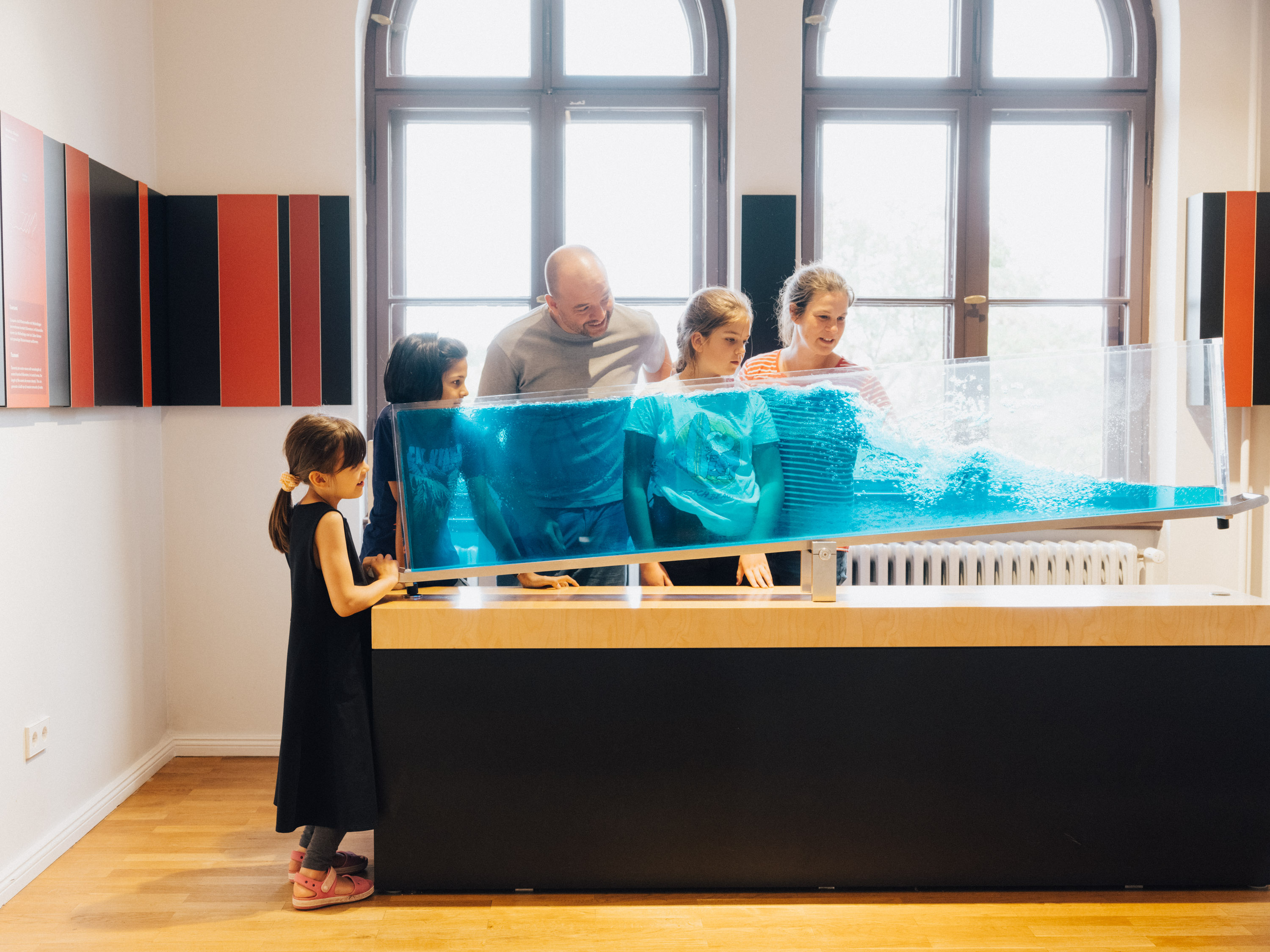 Eine Gruppe von Menschen steht um einen geneigten durchsichtigen Behälter herum, in dem eine blaue Flüssigkeit schwappt.