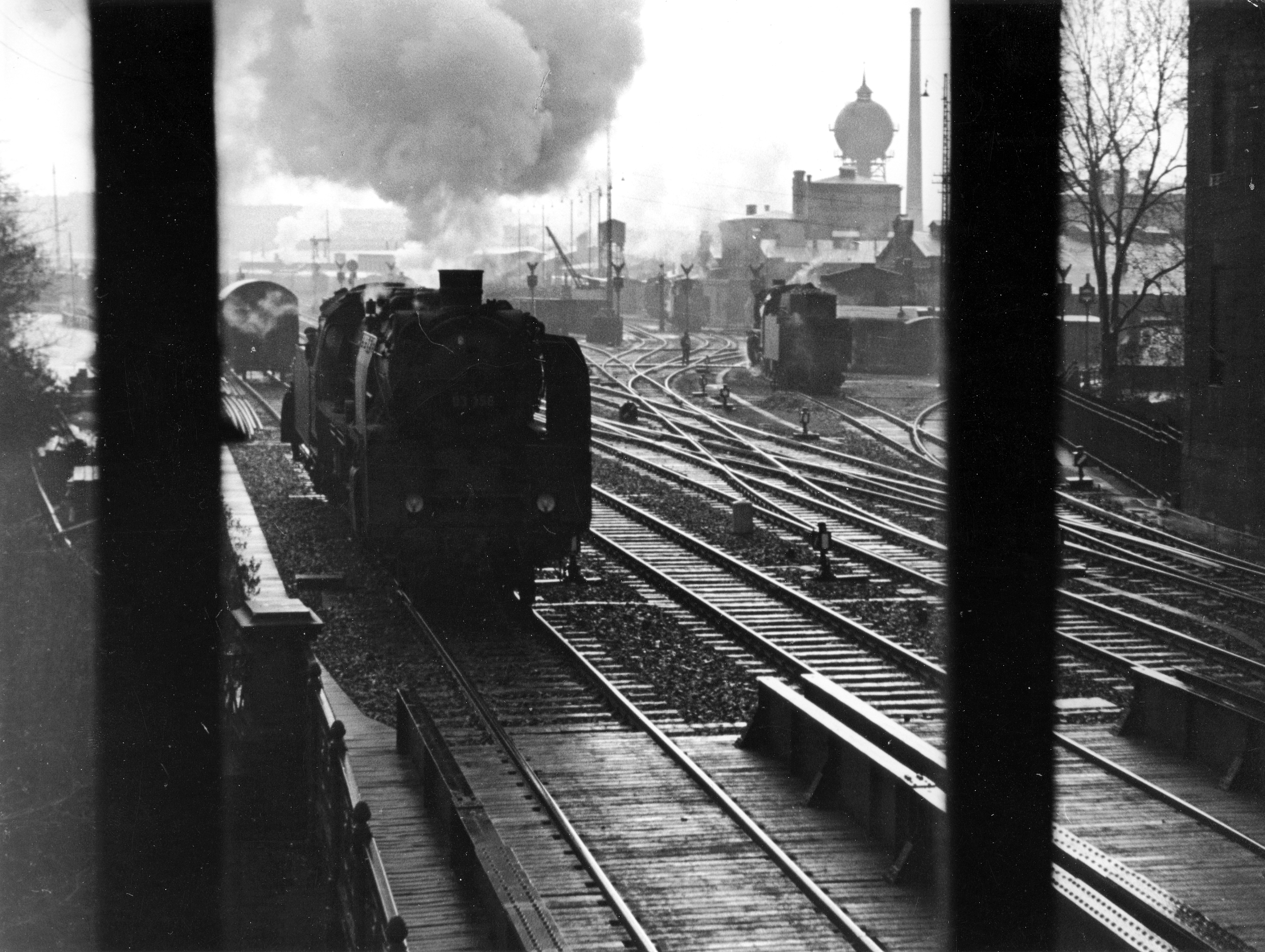 Schwarzweiß-Foto einer Dampflokomotive, die auf einer Schiene ins Bild fährt. Im Hintergrund sind ein Wasserturm und ein Lokschuppen zu erkennen.