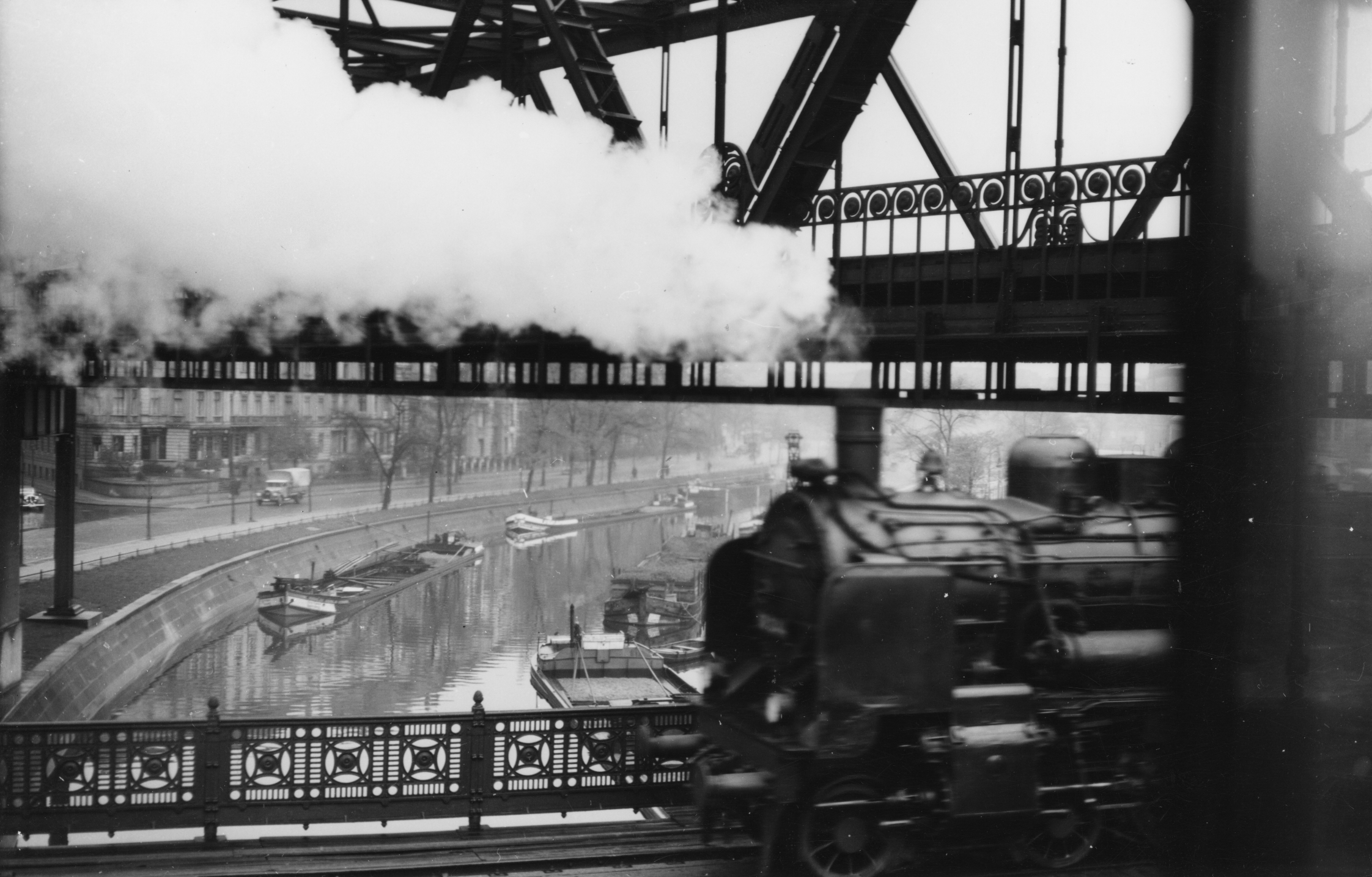 Schwarzweiß-Foto einer in Dampfwolken gehüllten Lokomotive, die auf einer Brücke einen Kanal überquert.