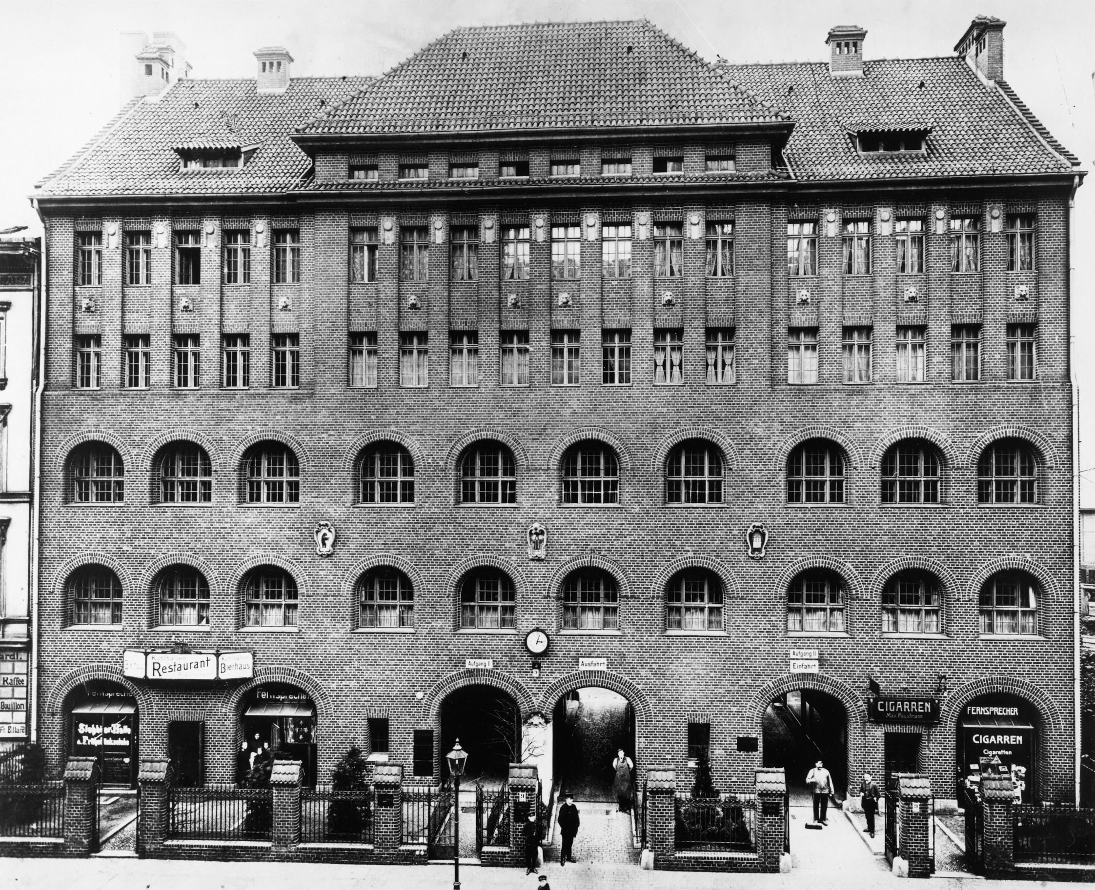 Schwarzweiß-Foto: Frontalansicht eines großen, vierstöckigen Backsteingebäudes. Im Erdgeschoss führen große Torbögen ins Innere.