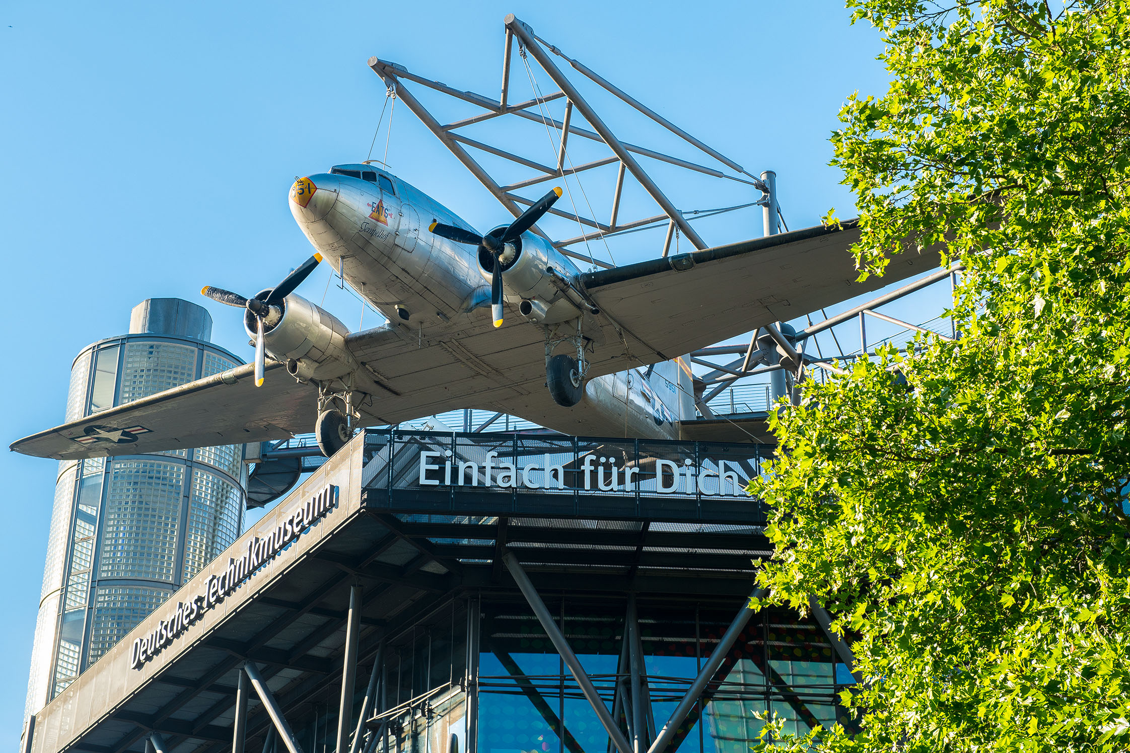 Ein Neubaugebäude mit Glasfassade. An einer Stahlkonstruktion auf dem Dach hängt ein Flugzeug. Unter dem Flugzeug hängt eine weiße Leuchtschrift „Deutsches Technikmuseum / Einfach für Dich“.