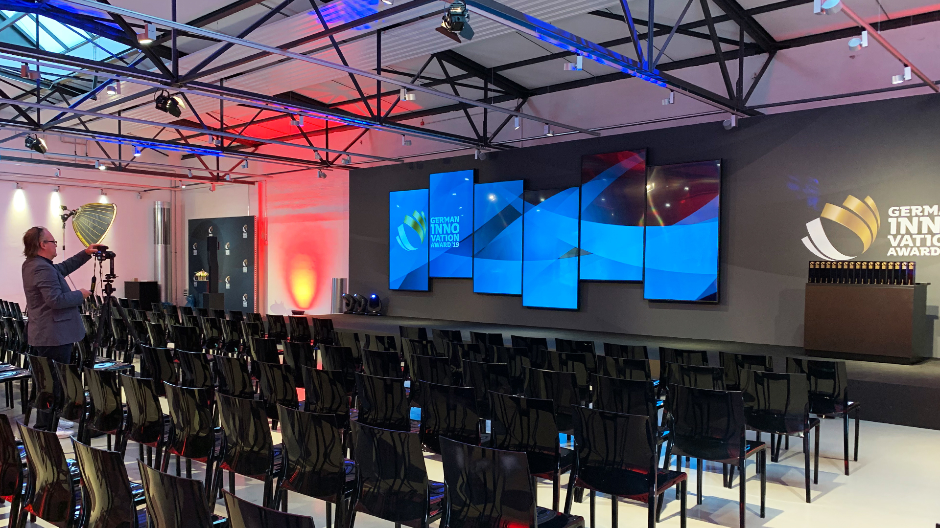 In einer Halle sind Stuhlreihen und ein Podium mit Monitoren an der Rückwand zu sehen.